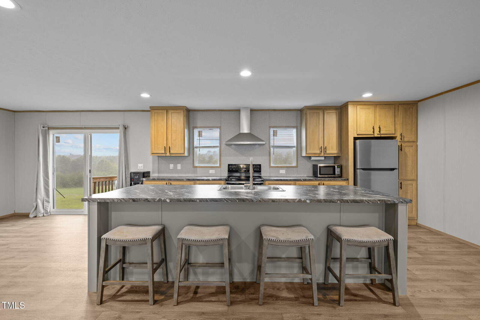 Modern kitchen with light wood cabinets, stainless steel appliances, island with marble countertop, four cushioned stools, and large windows letting in natural light. Sliding glass door opens to an outdoor view.