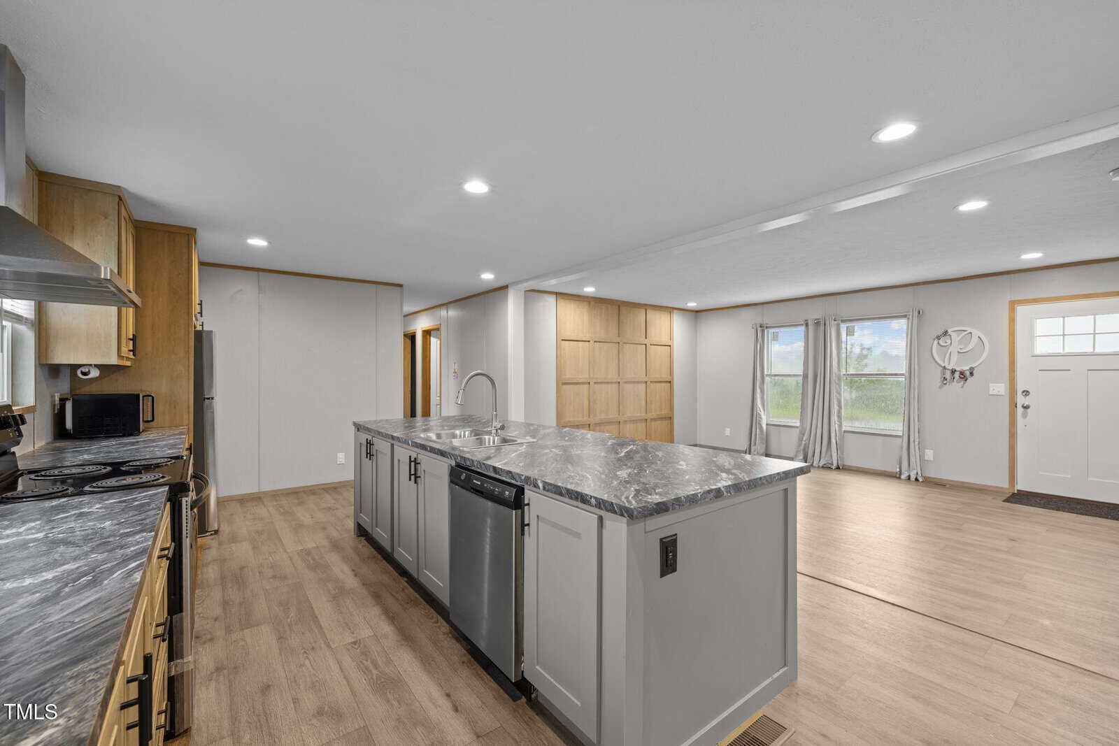 Spacious modern kitchen with light wood floors, large center island with marble countertop, stainless steel appliances, and open living area with big windows and front door visible in the background.