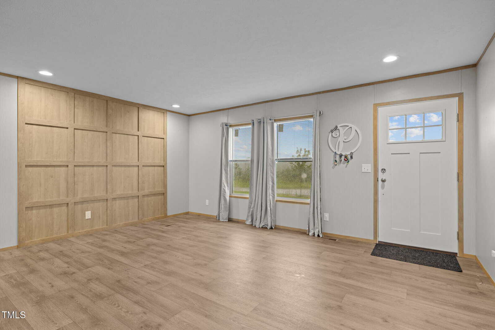Empty room with light wood floors, a grid-style accent wall, two windows with gray curtains, and a white front door with a mat. Small wall hooks are mounted near the door. Ceiling and walls are painted neutral colors.