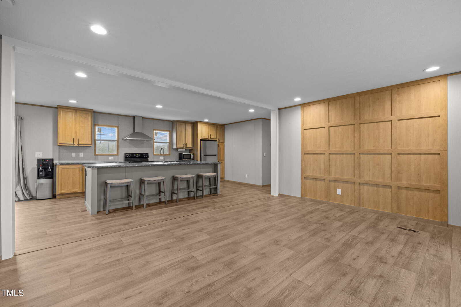 Open-plan room with light wood flooring, a kitchen area featuring an island with stools, stainless steel appliances, and wooden cabinets. The far wall has a large wooden accent panel. Recessed lighting is visible on the ceiling.