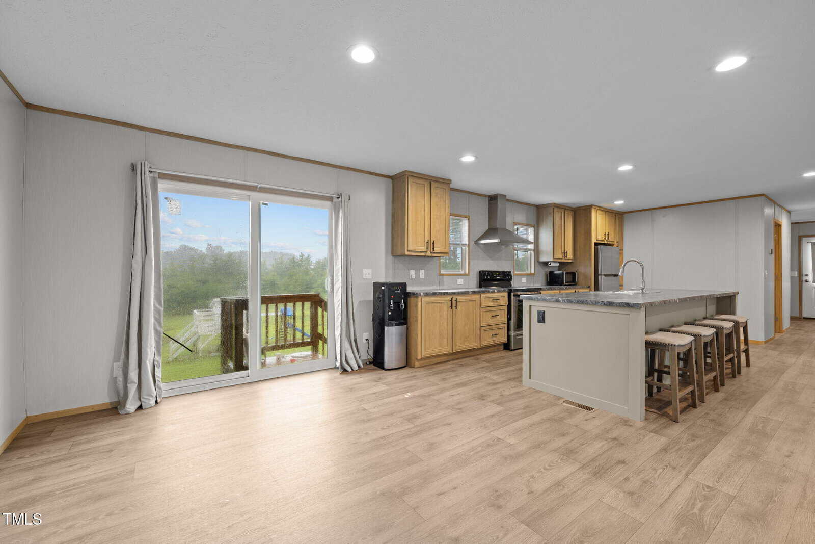 Modern kitchen with light wood floors, island with four stools, wood cabinets, stainless steel appliances, and sliding glass doors leading to a deck with a view of greenery outside. Room is brightly lit with recessed lighting.