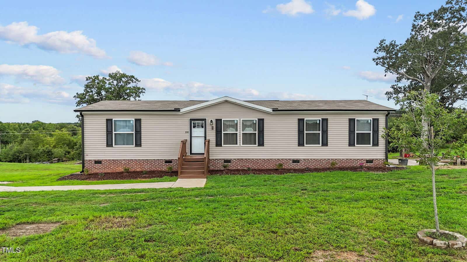 A single-story beige house with black shutters and a brick foundation sits on a large grassy lawn under a partly cloudy sky. Concrete steps lead up to the white front door. Trees and landscaping are visible in the background.