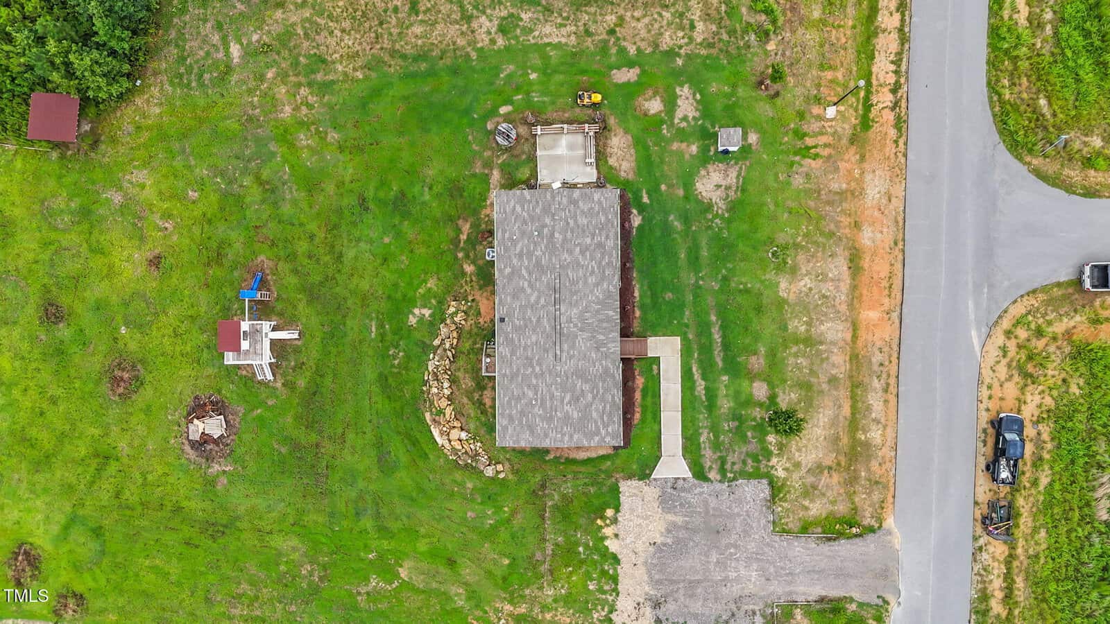 Aerial view of a house with a gray roof, surrounded by green grass, a driveway, and a nearby road. There's a small structure and fire pit in the yard, with scattered items on the lawn.