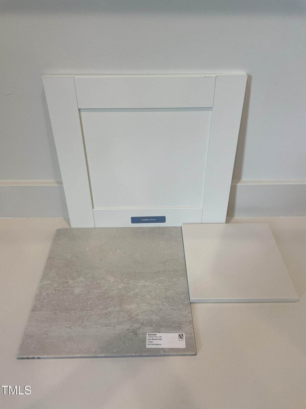 A white cabinet door sample stands upright against a light wall. In front of it, a large square gray tile and a smaller square white tile are placed on the floor. A small label is visible on the gray tile.