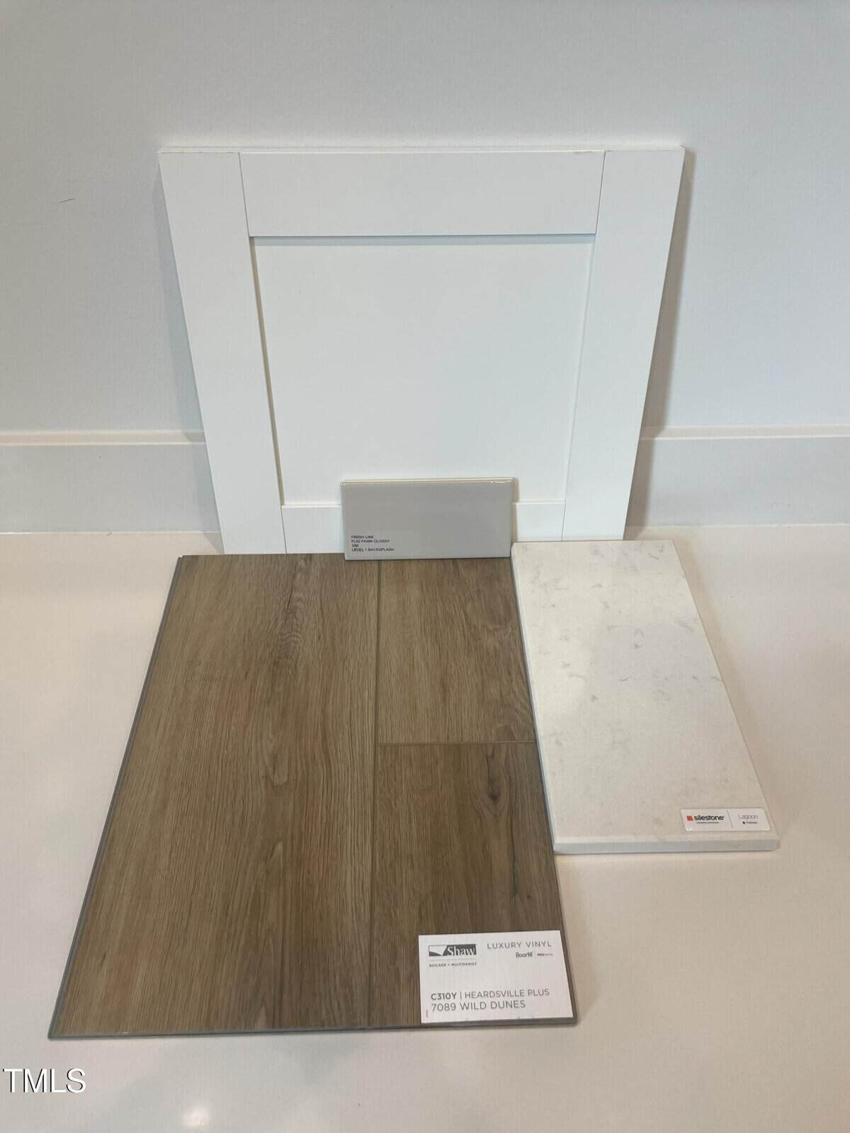 A white cabinet door, a white rectangular tile, a wood-look vinyl plank, and a white marble-patterned tile are displayed upright and on the floor against a white wall. Each item has a small label with text.