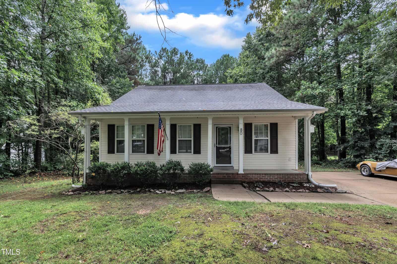 A small white house with a front porch, black shutters, and an American flag. The house is surrounded by trees, with a driveway on the right and a yellow car partially visible. The lawn is green and there are bushes by the porch.