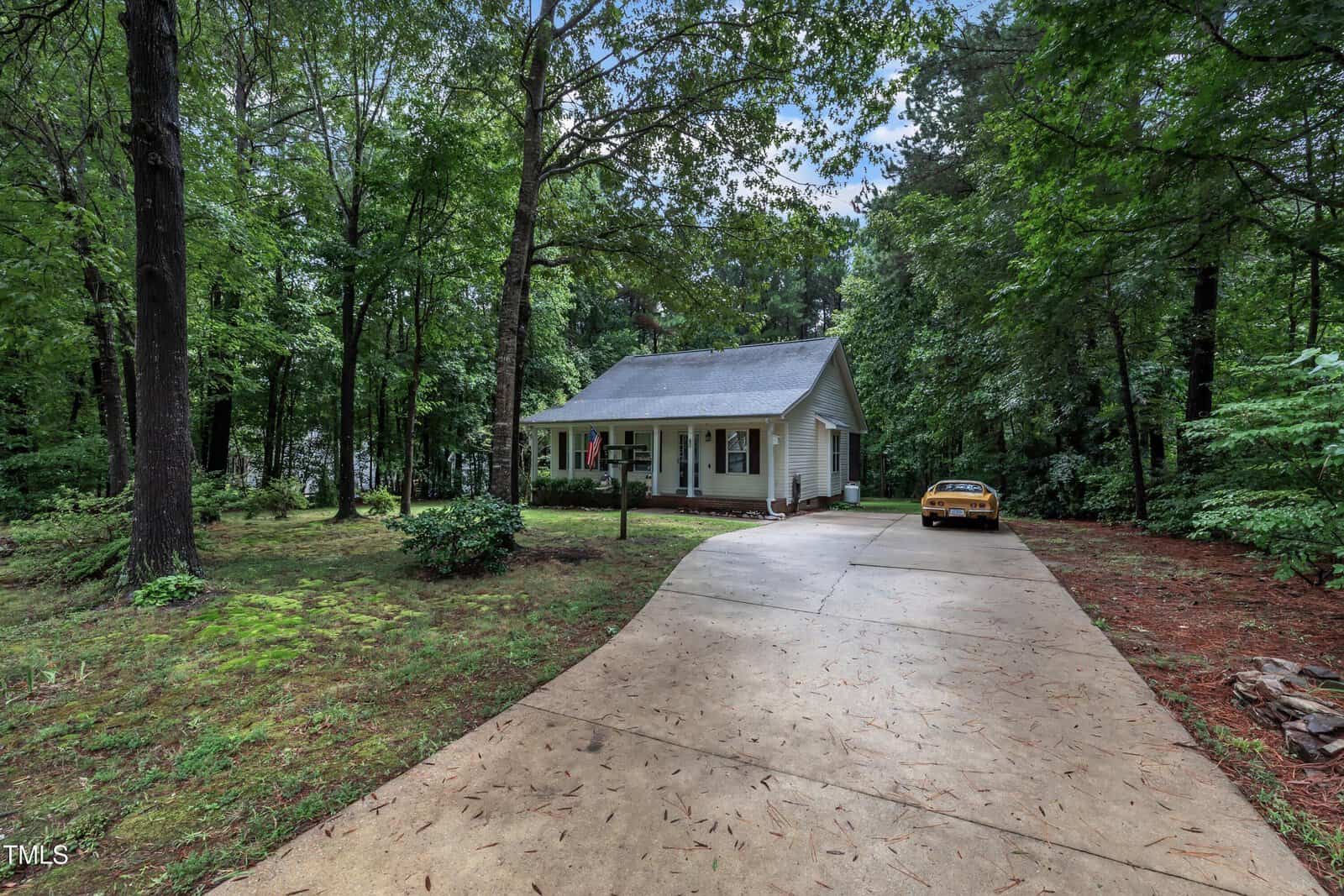 A small white house with a front porch sits among tall trees. A concrete driveway leads to the house, with a yellow car parked near the entrance. The yard is grassy and surrounded by lush greenery.