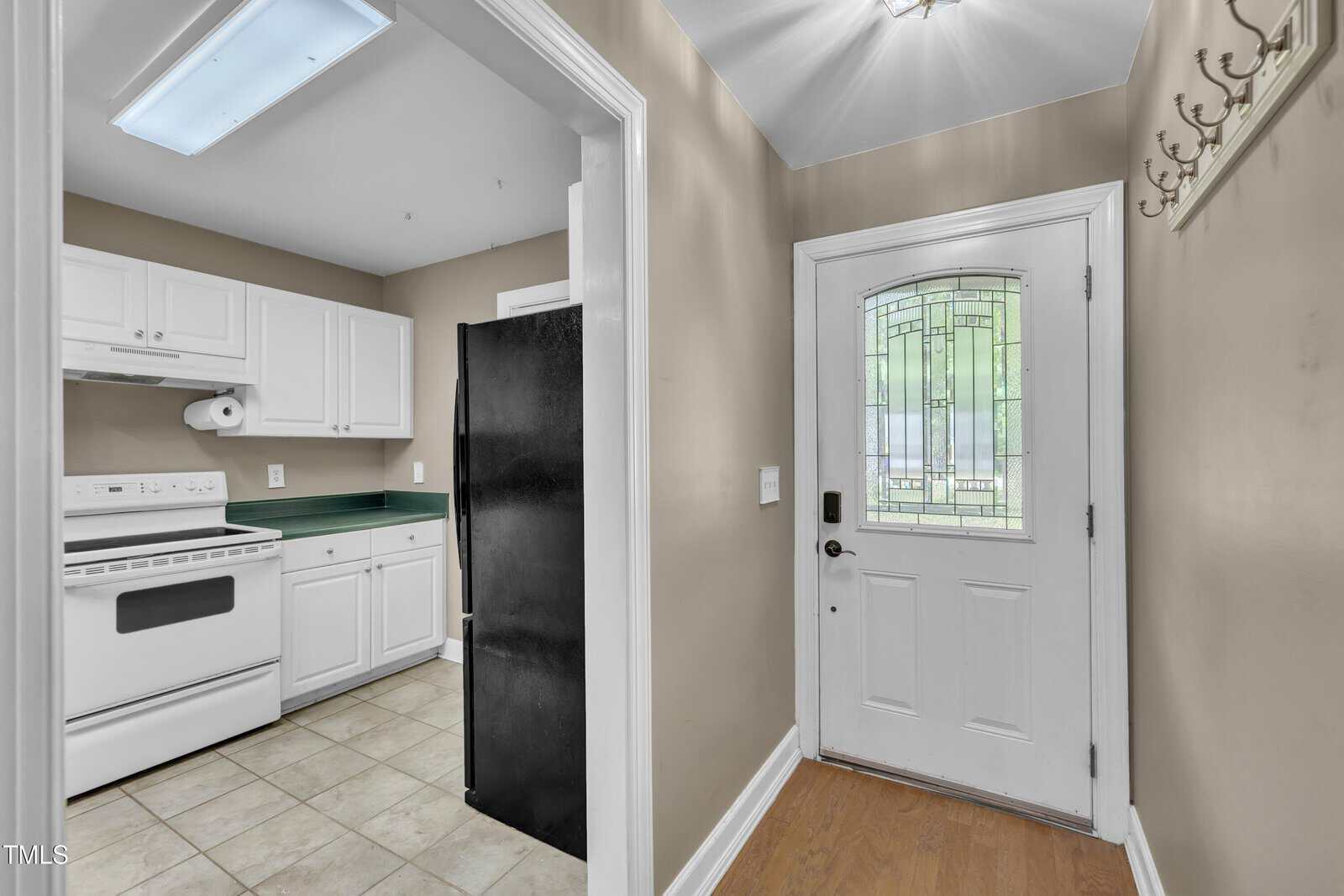 A small entryway with a white door featuring decorative glass, next to a wall with hooks. The entry opens into a kitchen with white cabinets, a white stove, and a black refrigerator. The floor is tiled in the kitchen area.