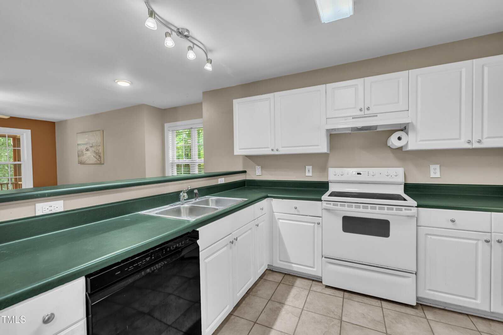 A kitchen with green countertops, white cabinets, a white stove and oven, a dishwasher, and a double sink. The room has beige walls, tiled flooring, and a window letting in natural light.