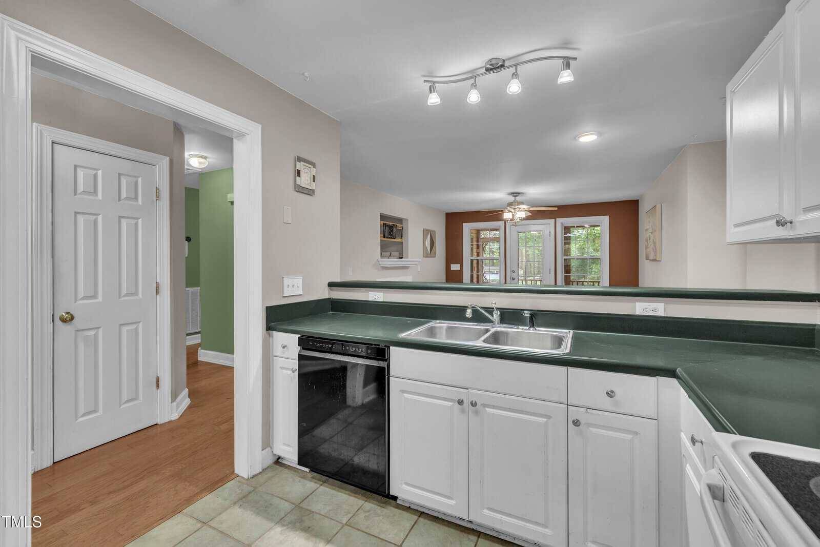 Modern kitchen with white cabinets, green countertops, double sink, black dishwasher, and tiled floor; view into a dining area with wooden floor and glass doors leading outside.