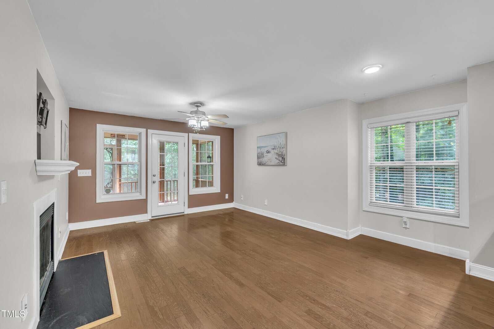Unfurnished living room with hardwood floors, a ceiling fan, a fireplace, two windows with blinds, a glass door leading to a deck, and light brown and white painted walls. A framed picture hangs on one wall.