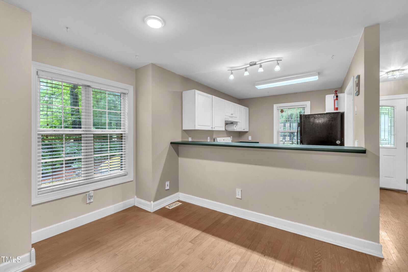 A small kitchen with white cabinets, black appliances, and a green countertop, seen from an adjacent dining area with light brown wood floors and a window with white blinds. A door is visible in the background.