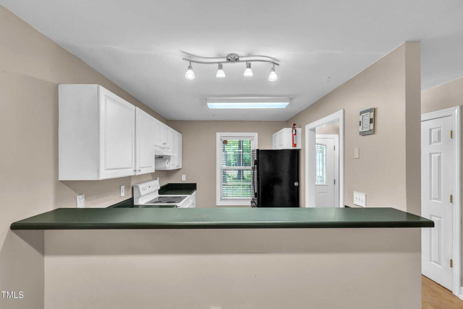 A kitchen with white cabinets, green countertops, a black refrigerator, a white stove, and a window with blinds. Overhead lighting and a fire extinguisher are visible. The room has beige walls and a pass-through counter.