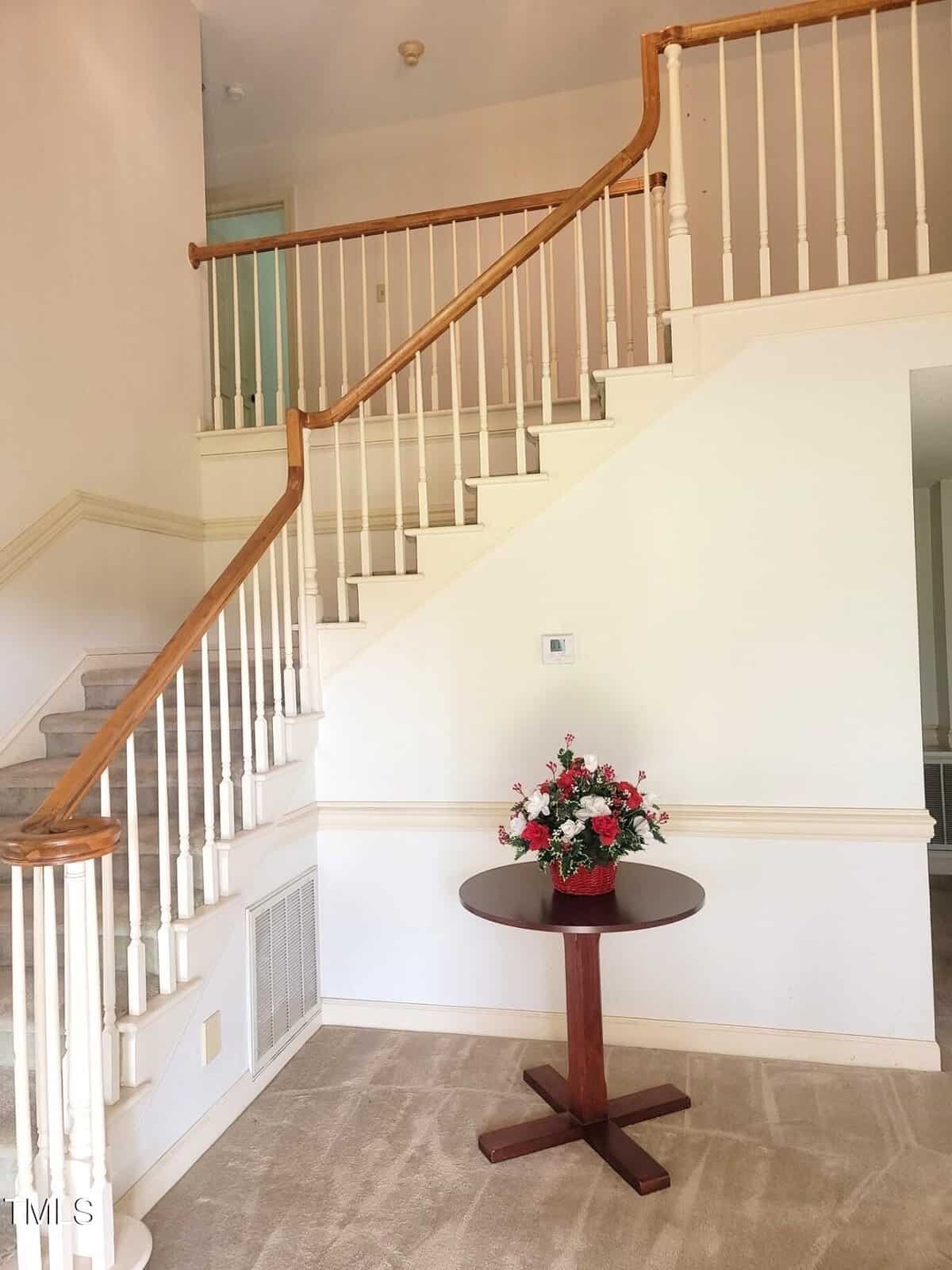 A small wooden round table with a floral arrangement sits below a carpeted staircase with white spindles and a wooden handrail in a bright, neutral-toned entryway.