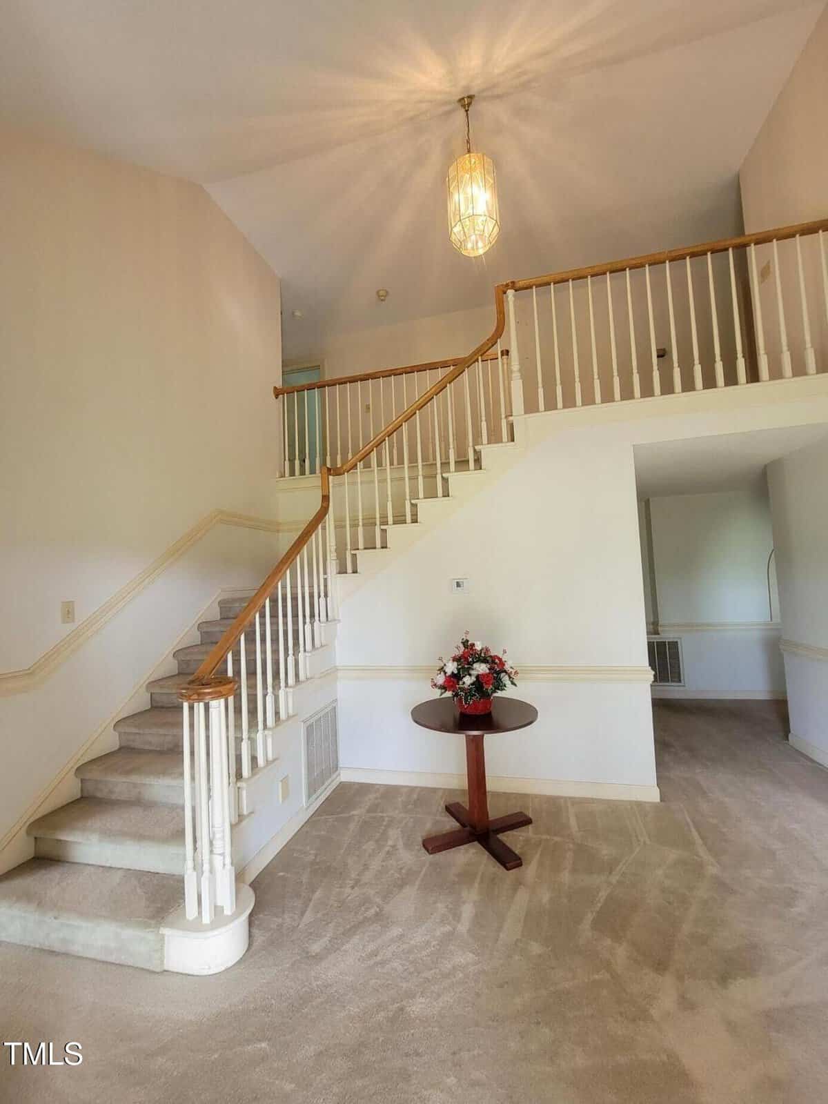 A carpeted staircase with white railings leads to an upper floor in a spacious room with beige walls and carpet. A round wooden table with a flower arrangement sits at the base of the stairs under a hanging light fixture.