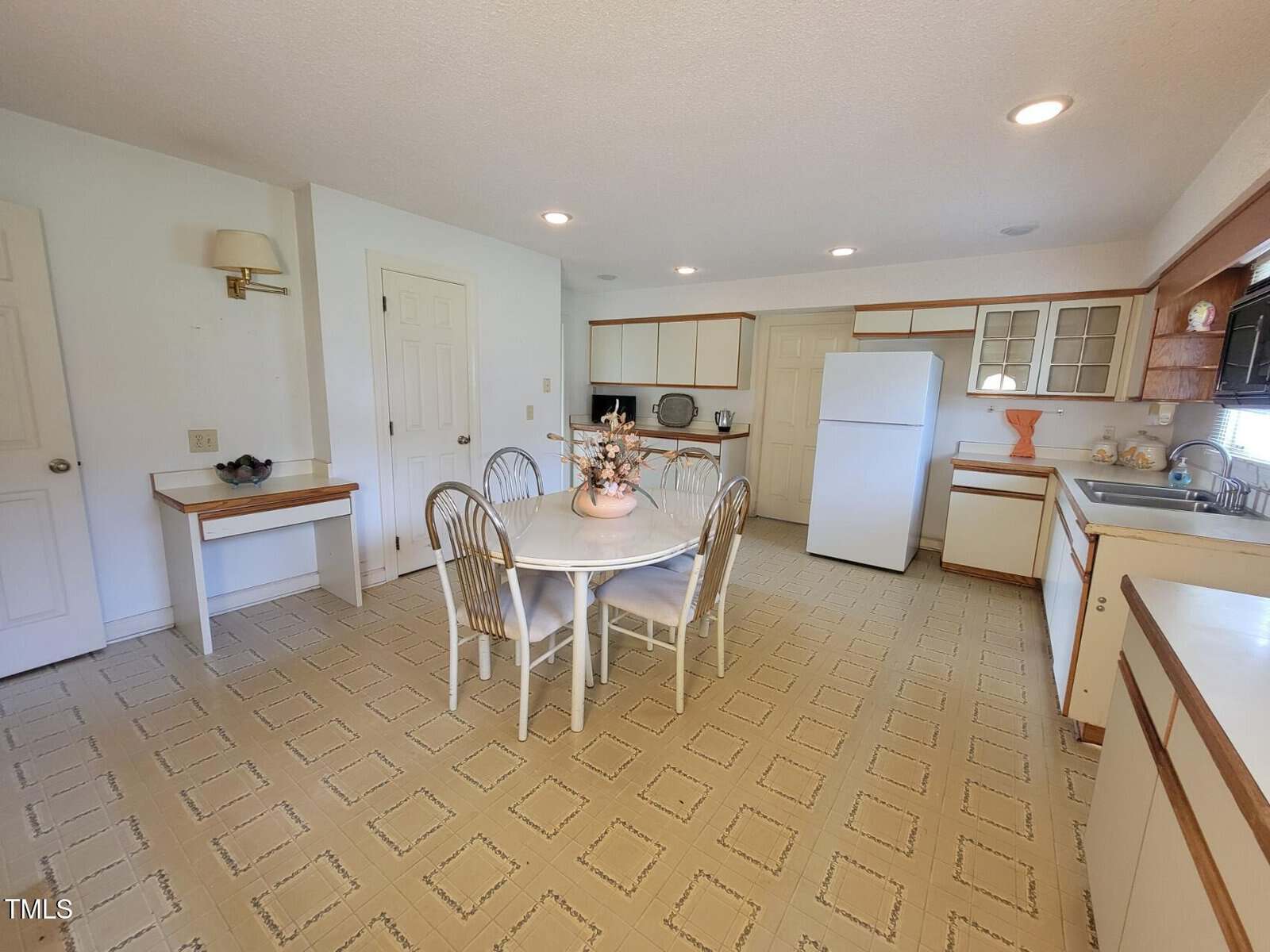 A kitchen with light-colored cabinets, a white refrigerator, stove, and microwave. A square dining table with four chairs is in the center, decorated with a flower arrangement. The floor has a patterned linoleum.