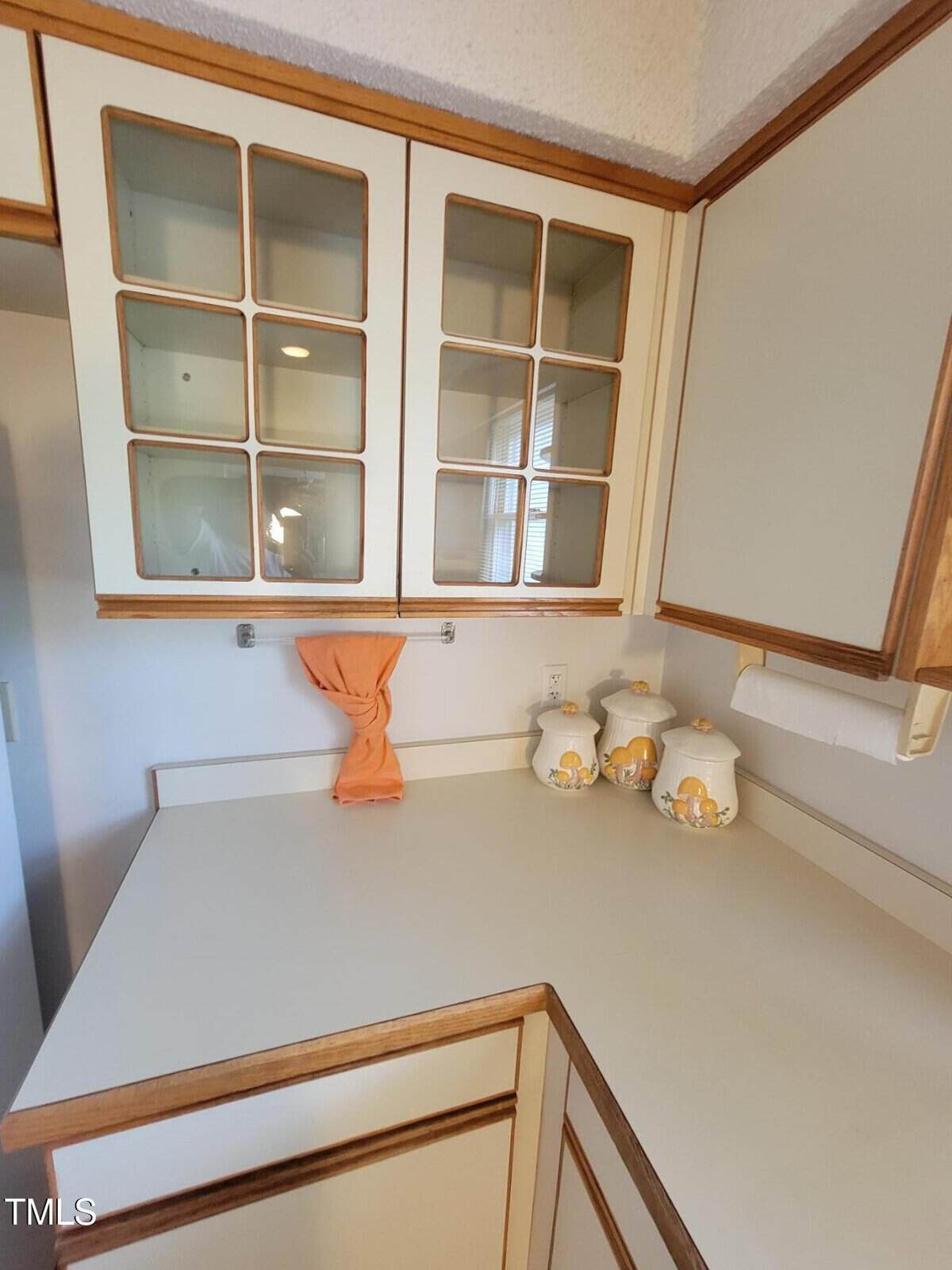 A kitchen counter with cream cabinets and wooden trim, featuring three decorative ceramic canisters with fruit designs, a paper towel holder, and a peach-colored dish towel hanging below glass-front cabinets.