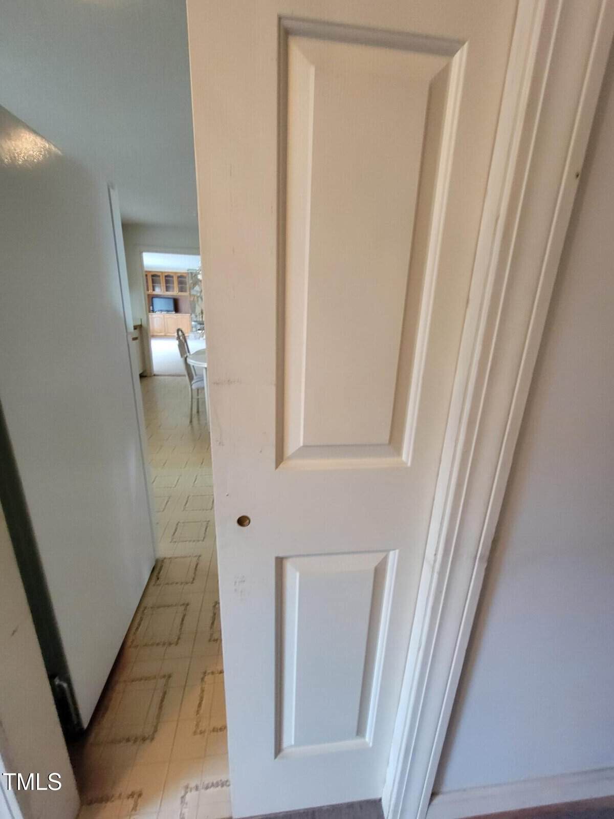 A partially open white paneled door with a small hole near the handle area reveals a view into a kitchen with light-colored flooring and visible cabinetry in the background.