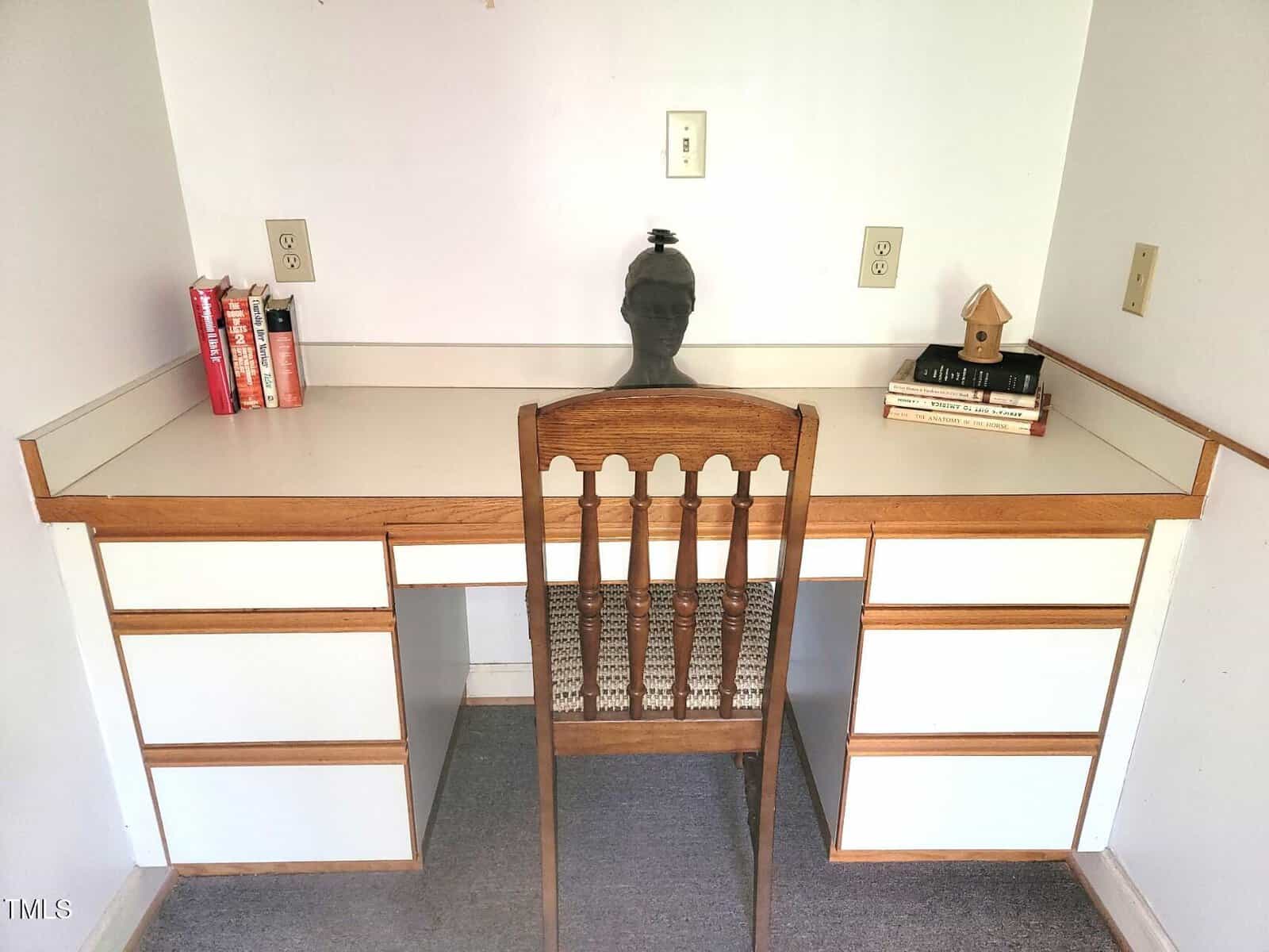 A wooden desk with white drawers, a wooden chair, a few books on both ends of the desk, a small birdhouse, and a bust sculpture placed in the center against a white wall with several electrical outlets.