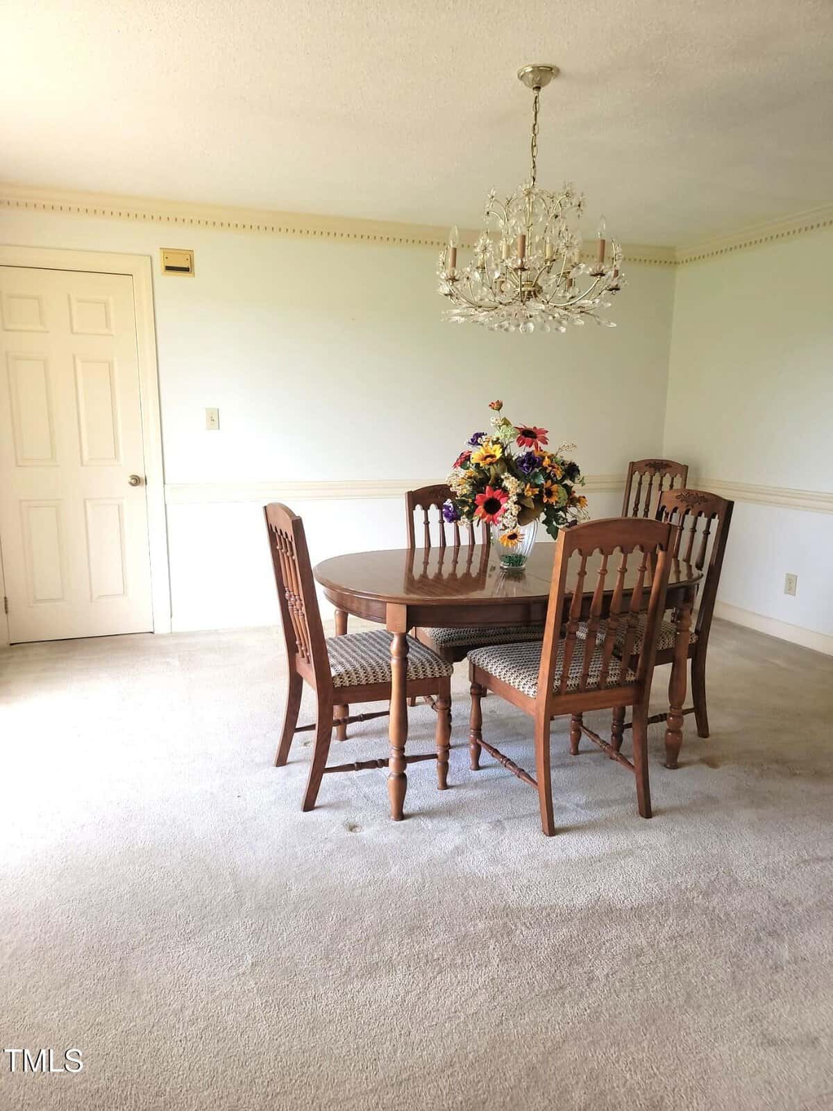 A dining room with a round wooden table and four matching chairs, a vase of colorful flowers on the table, a chandelier overhead, and a closed door in the background. The room has carpeted floors and white walls.