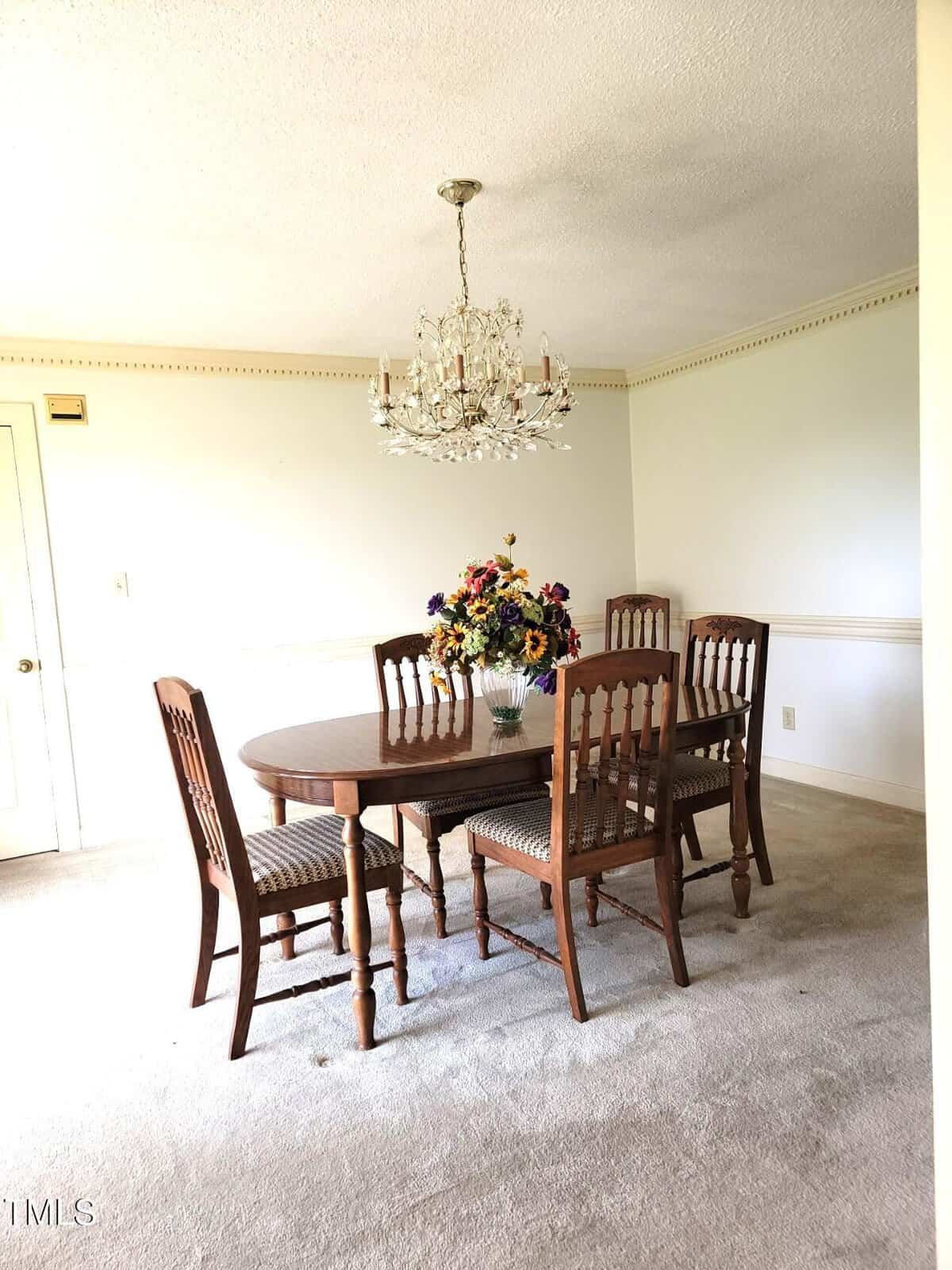 A dining room with a wooden table, six matching chairs, a vase of colorful artificial flowers as a centerpiece, a crystal chandelier above, and light-colored carpeted flooring. The walls are light with decorative trim.