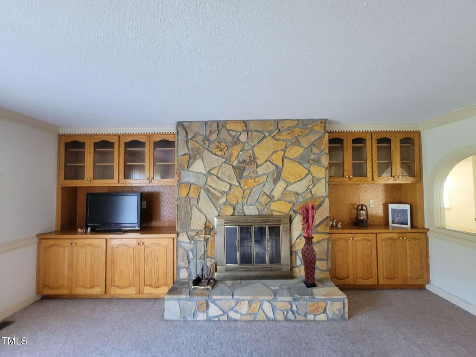A stone fireplace is centered between built-in wooden cabinets with glass doors; one side has a TV, the other has vases and decor. The room has carpet and a neutral color scheme.