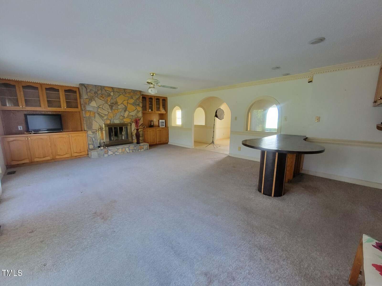 Spacious living room with built-in wood cabinets, a stone fireplace, beige carpet, ceiling fan, round table, and arched doorways leading to another area; light streaming through windows.