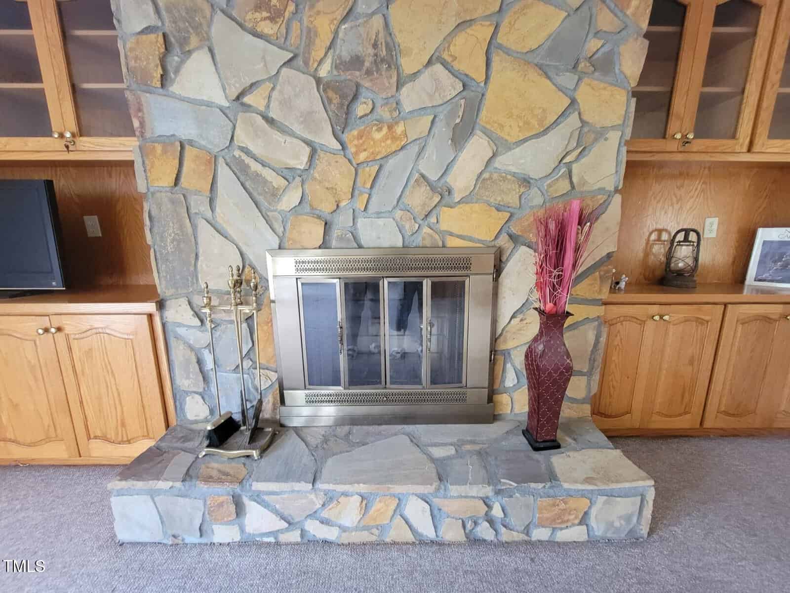 Stone fireplace with a metal screen, flanked by light wood cabinets. On the left is a TV and fireplace tools; on the right, a tall red vase with decorative sticks and a small basket. Carpeted floor in front.