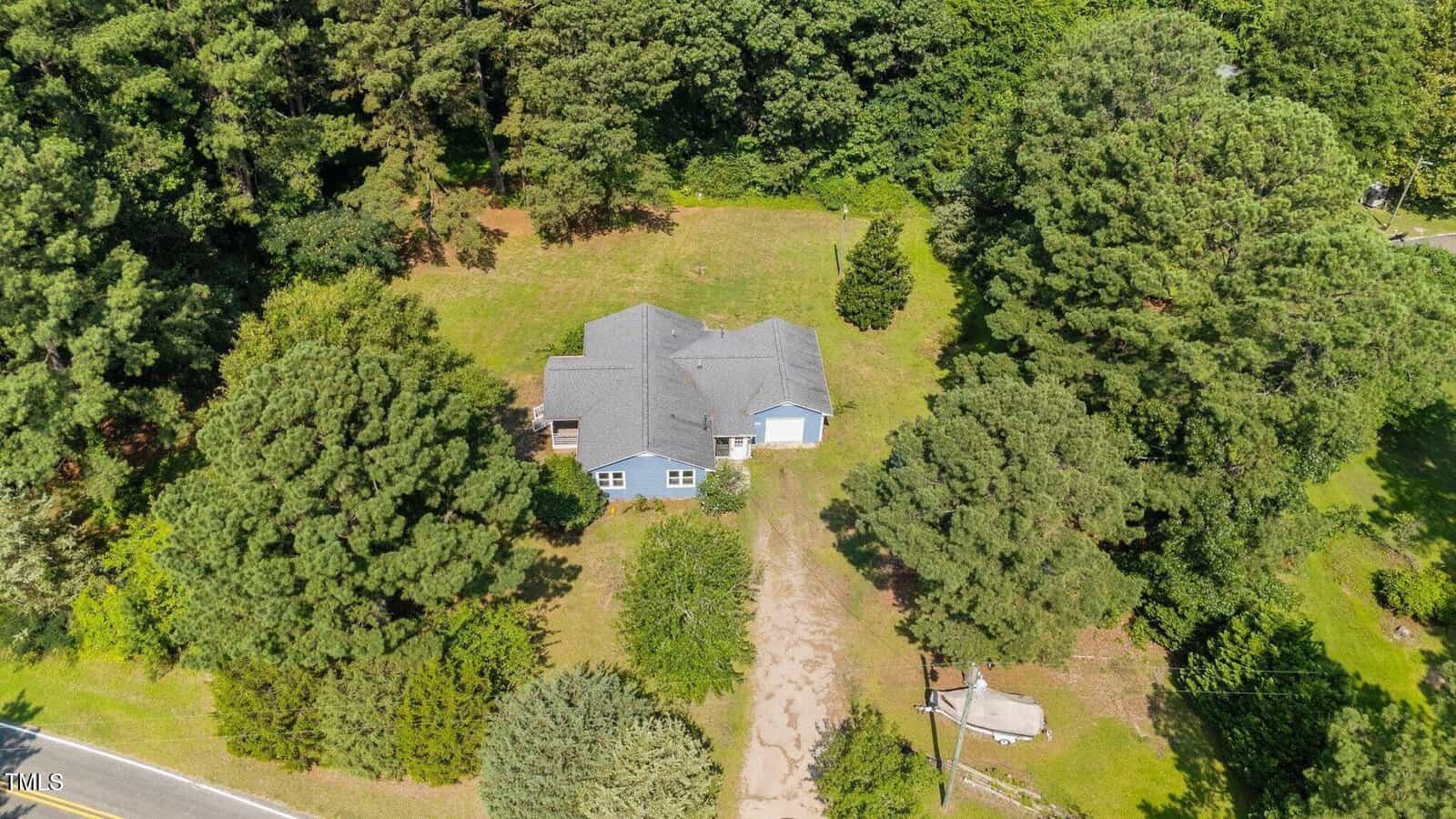 Aerial view of a single-story house with a gray roof surrounded by large green trees and grass, with a dirt driveway leading to the front from a nearby road.