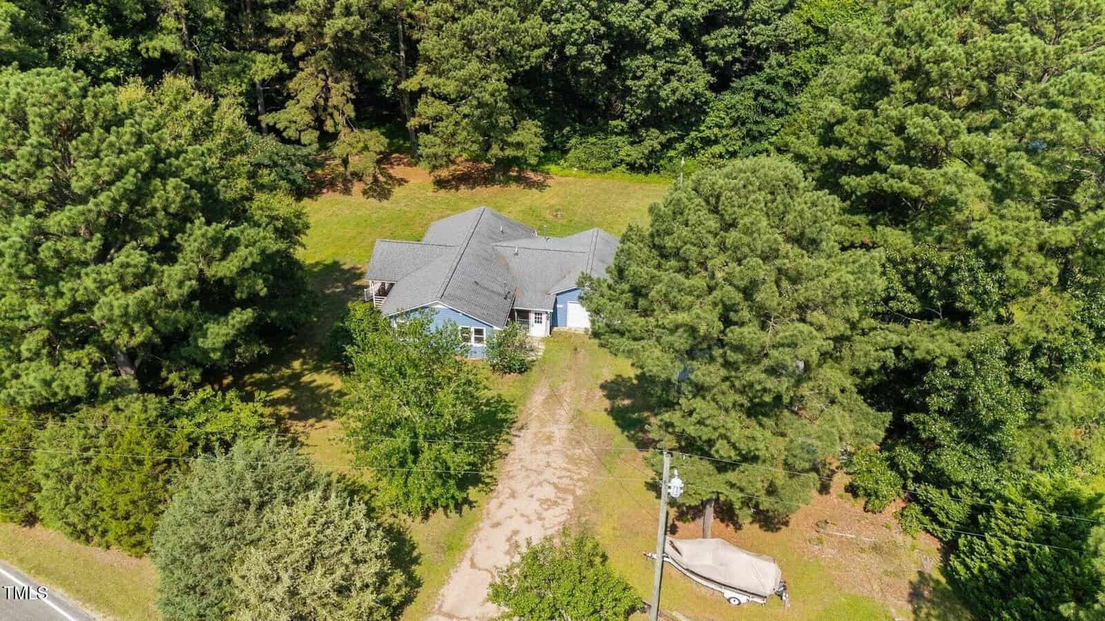 Aerial view of a blue house with a gray roof surrounded by tall trees and greenery. A dirt driveway leads to the house, with a covered boat parked beside it. Dense forest is visible in the background.