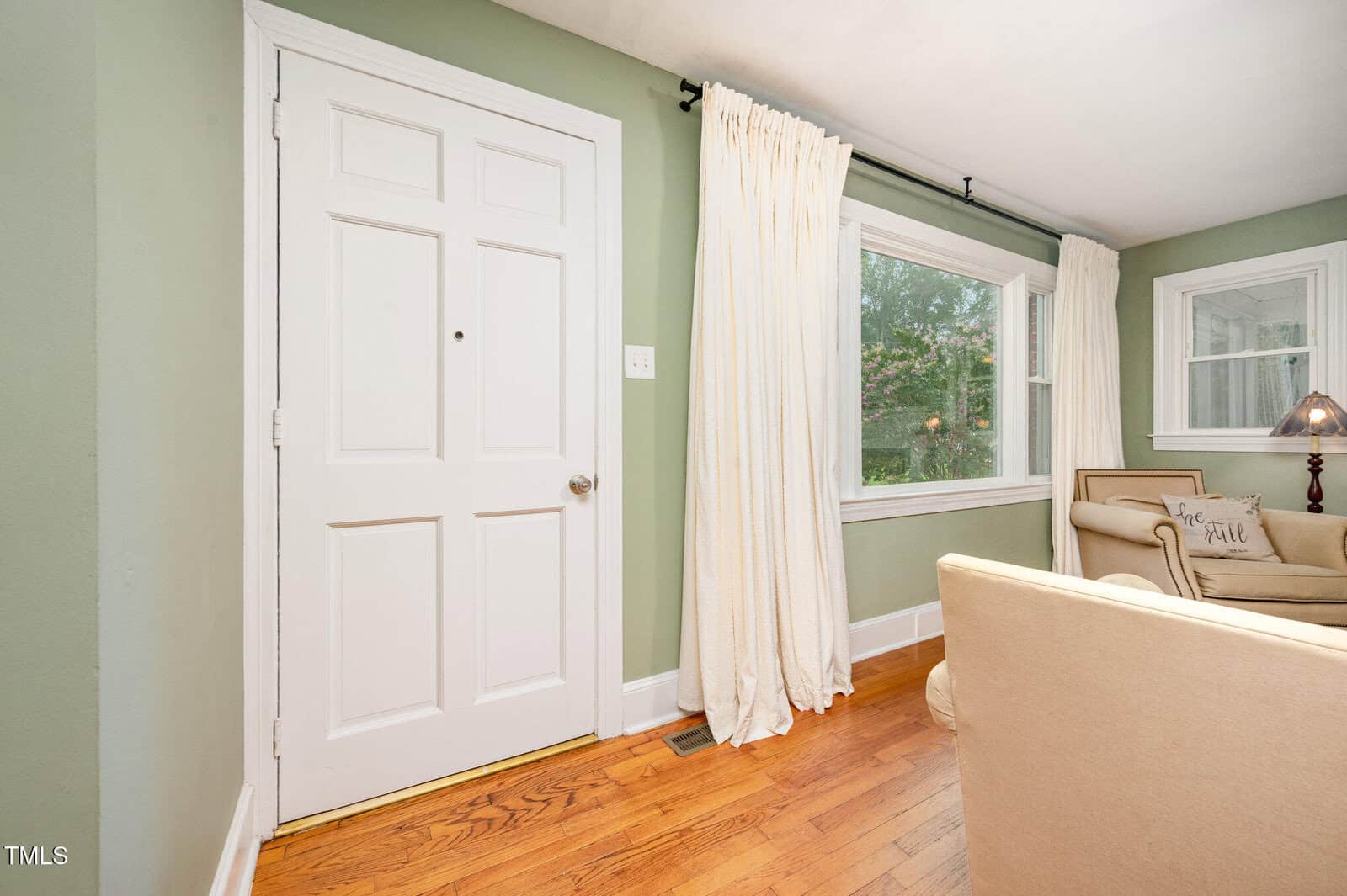 A white door on a light green wall is next to a window with white curtains. Hardwood floors and beige armchairs are visible, along with a side table and lamp by another window in the living room.