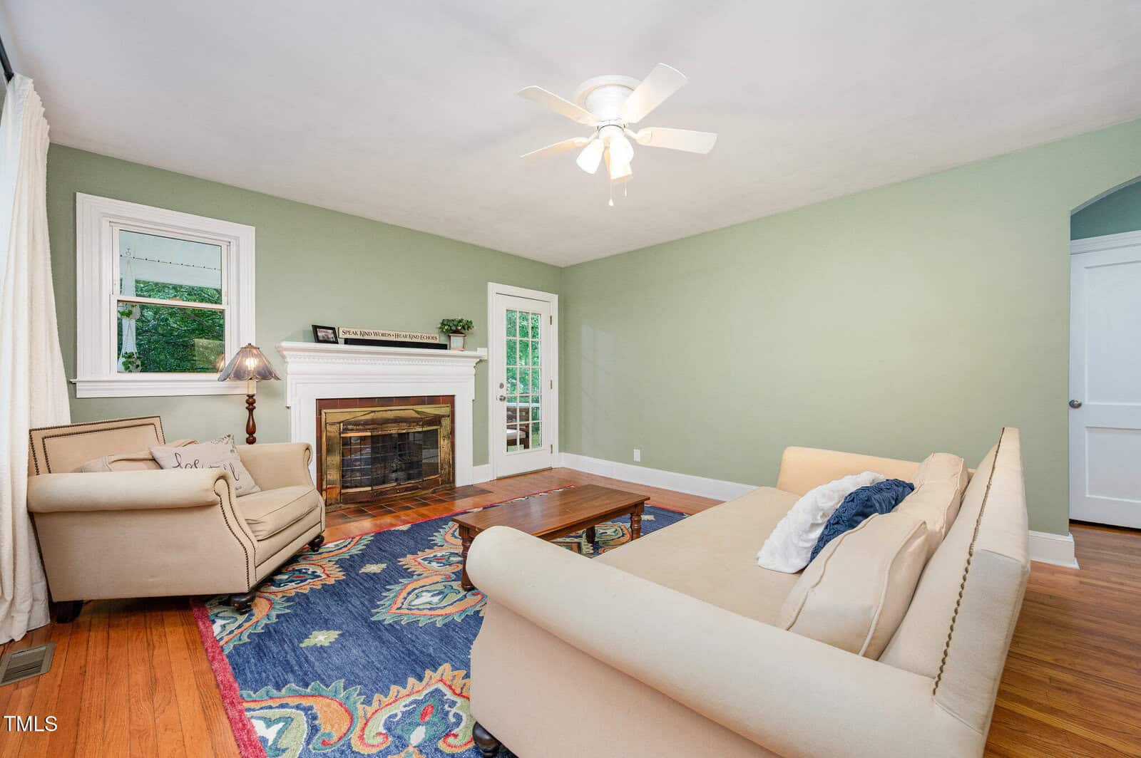 A living room with green walls, hardwood flooring, a fireplace with a wooden mantel, a beige sofa, an armchair, a wooden coffee table, a colorful rug, and a ceiling fan with lights.