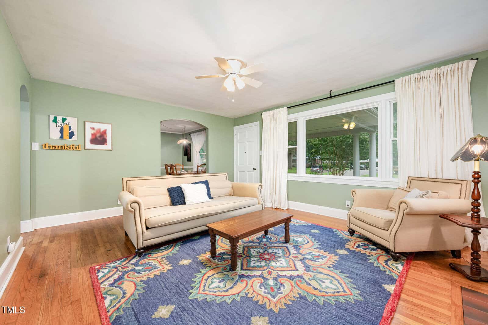 A living room with light green walls, a ceiling fan, a large window with white curtains, two beige sofas, a wooden coffee table, a patterned area rug, and framed artwork on the wall.