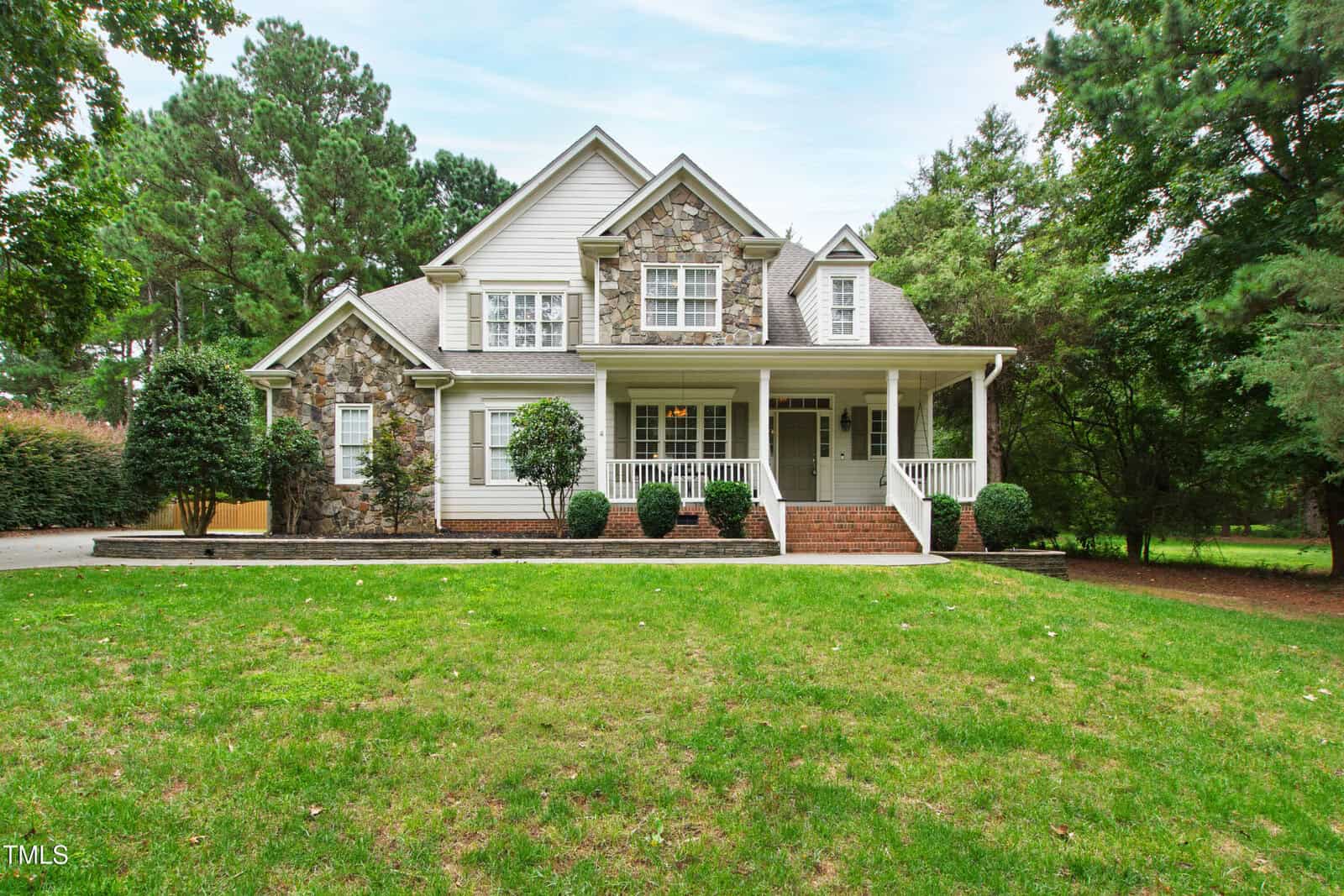 Charming two-story house at 145 Traylee Drive, Wake Forest, featuring a stone and siding exterior, front porch with white columns, brick steps, and manicured bushes. Surrounded by a lush green lawn and tall trees, with a driveway visible on the left. The sky is partly cloudy.