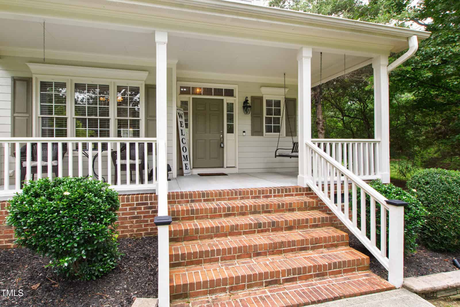 Front porch of a house with white railings, brick steps, gray door, and a “Welcome” sign. Two windows, a porch swing, bushes, and trees are visible. The porch has white columns and a light fixture by the door.