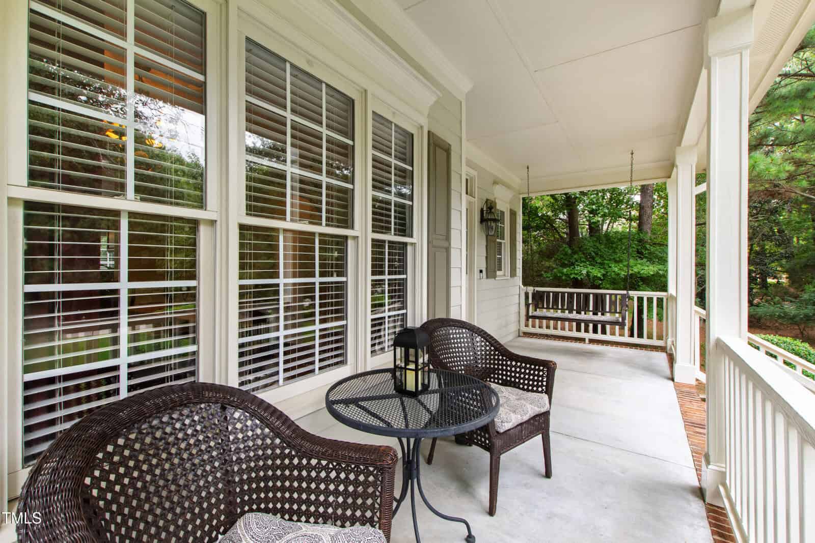 A covered porch features two wicker chairs and a round table on a concrete floor. A porch swing hangs at one end. White railings and window blinds face a green, wooded yard.