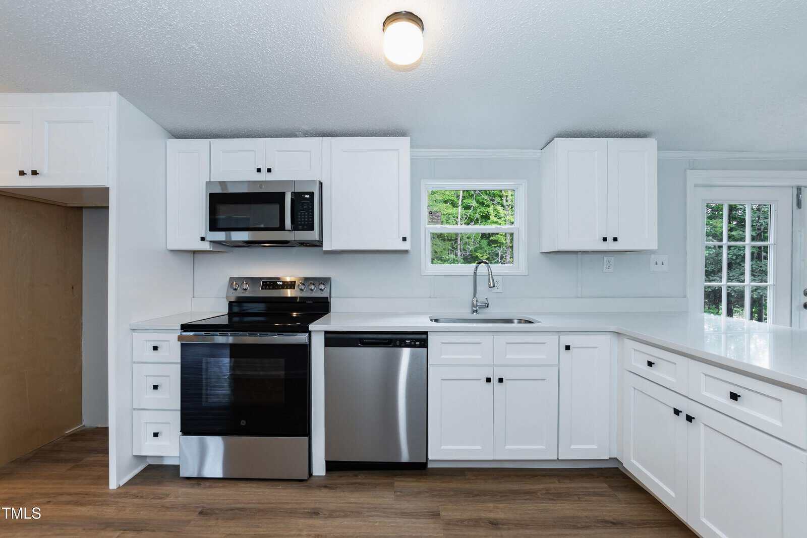 Modern kitchen with white cabinets, stainless steel appliances including a stove, microwave, and dishwasher, a single sink below a window, white countertops, and wood flooring. A door with glass panes is visible on the right.