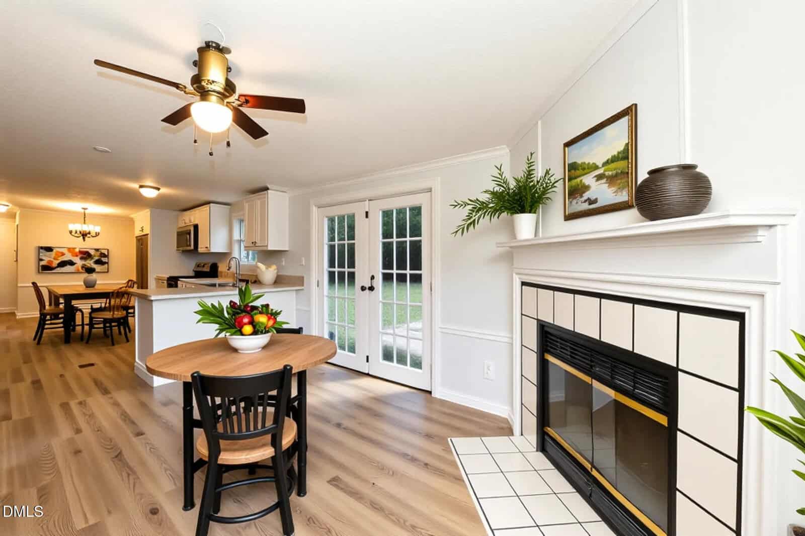 A dining area with a round wooden table and chairs, a ceiling fan above, French doors leading outside, a kitchen in the background, and a fireplace with art and a black vase on the mantel.