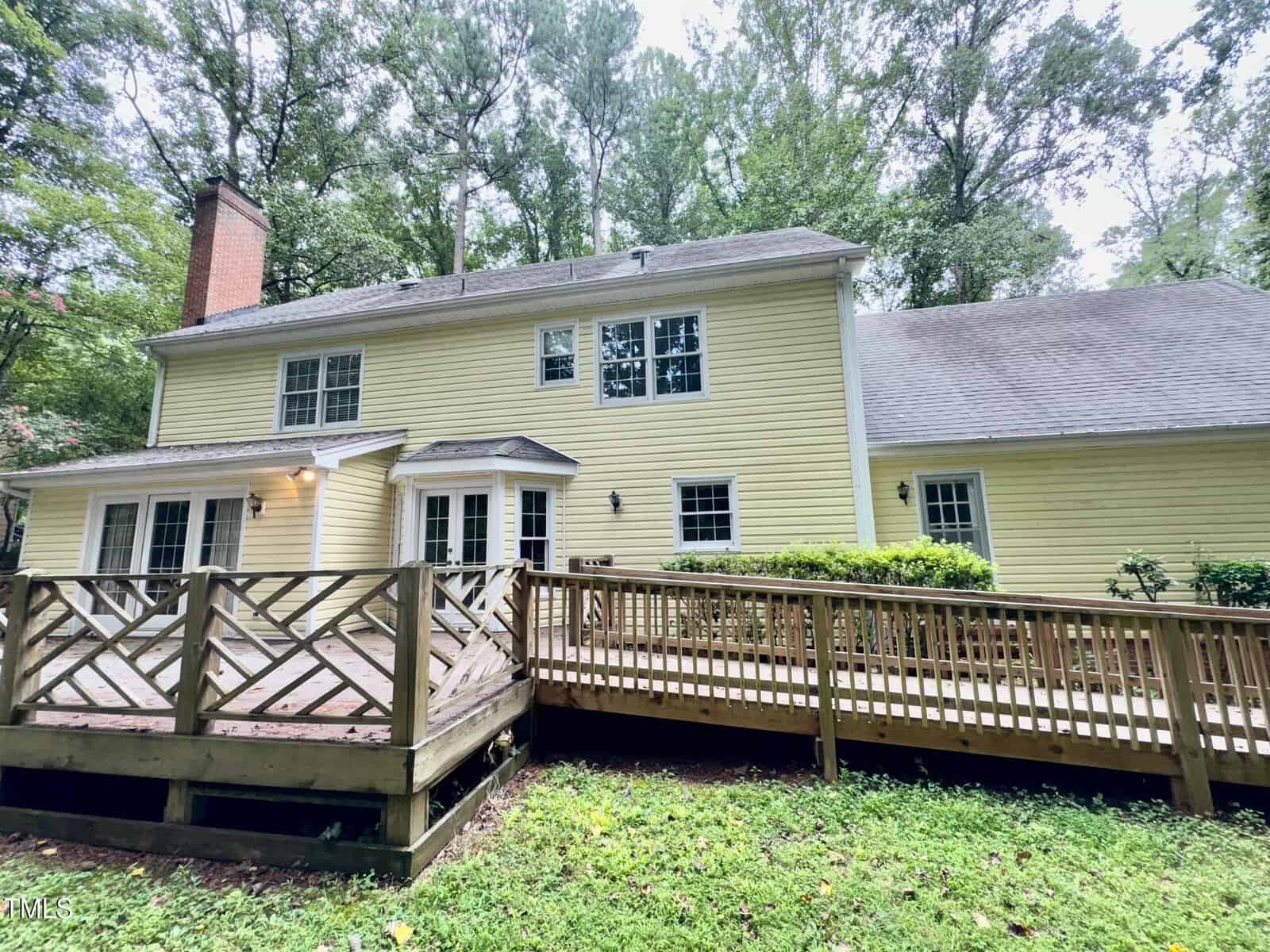 Two-story yellow house with white trim, a shingled roof, brick chimney, and several windows. A wooden deck with a ramp extends from the back, surrounded by grass and trees.