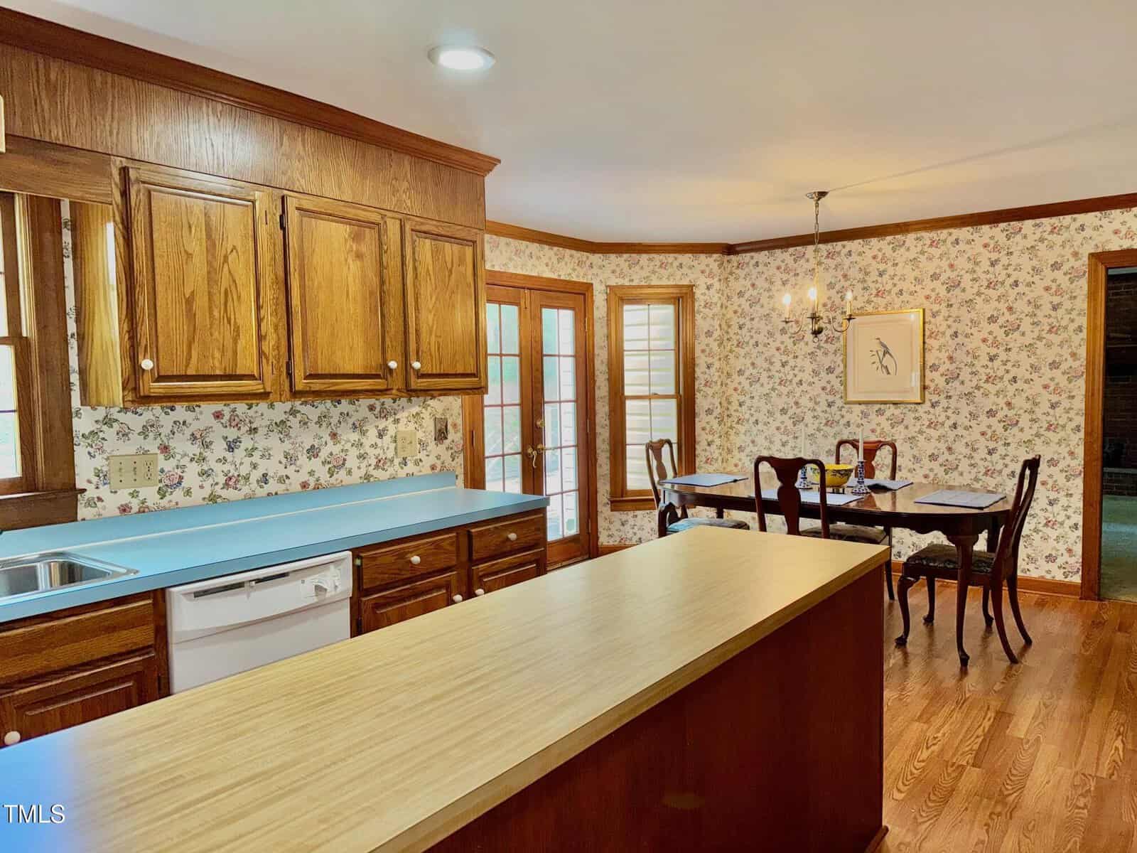 A kitchen with wood cabinets, a light-colored countertop island, blue countertops, a dishwasher, and a sink. The adjacent dining area features a table with chairs, a chandelier, floral wallpaper, and double French doors.