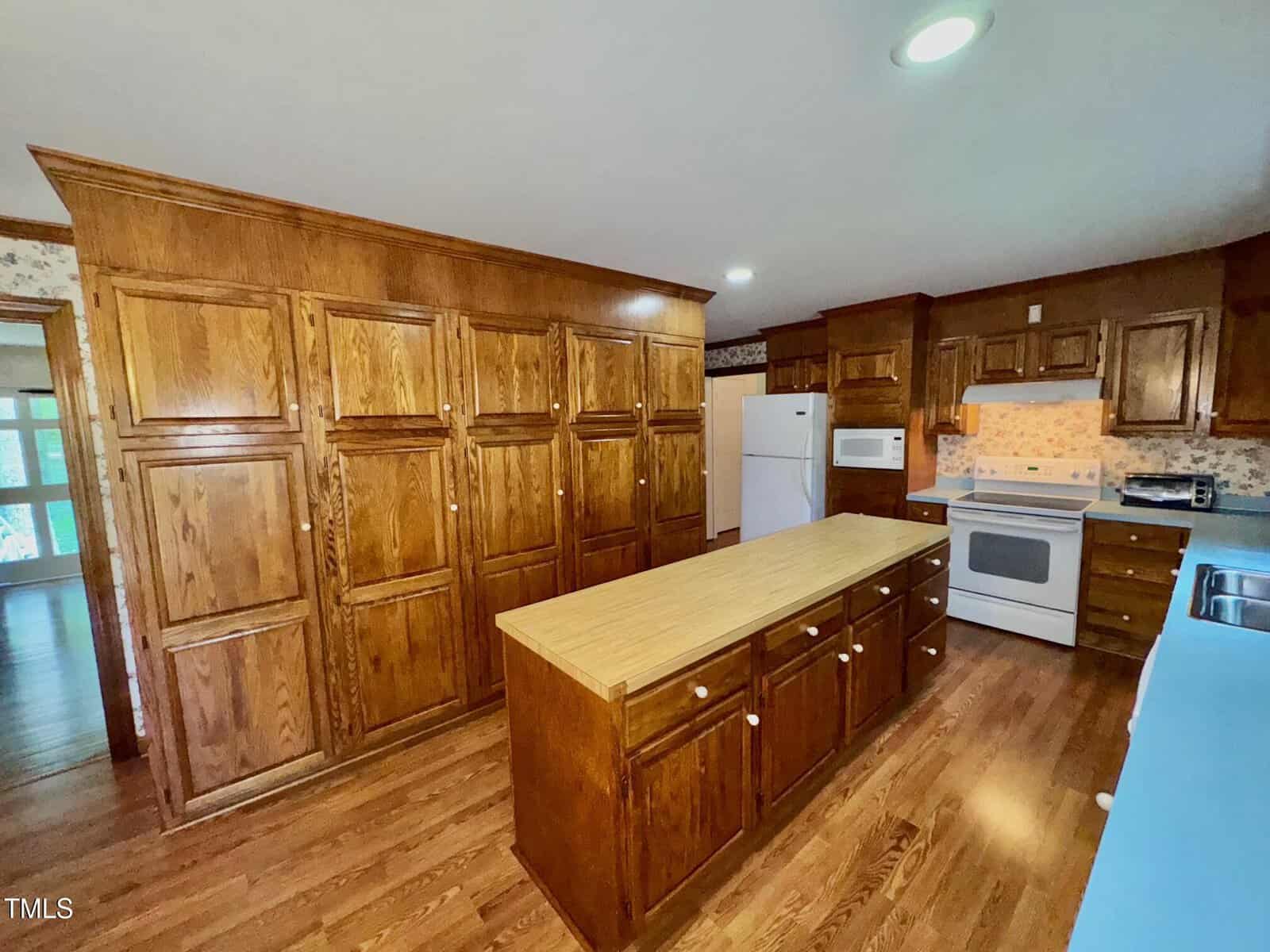 Spacious kitchen with wooden cabinets, a large central island with drawers, white appliances including a stove, microwave, refrigerator, and dishwasher, wood flooring, and overhead lighting.