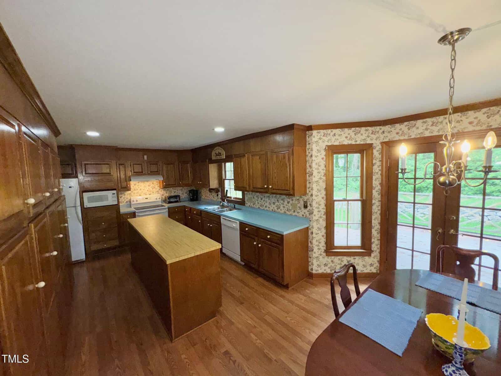 A spacious kitchen with wood cabinets, an island, white appliances, and light countertops, adjacent to a dining area with a round table, chandelier, and double glass doors leading outside. Wood floors and floral wallpaper throughout.