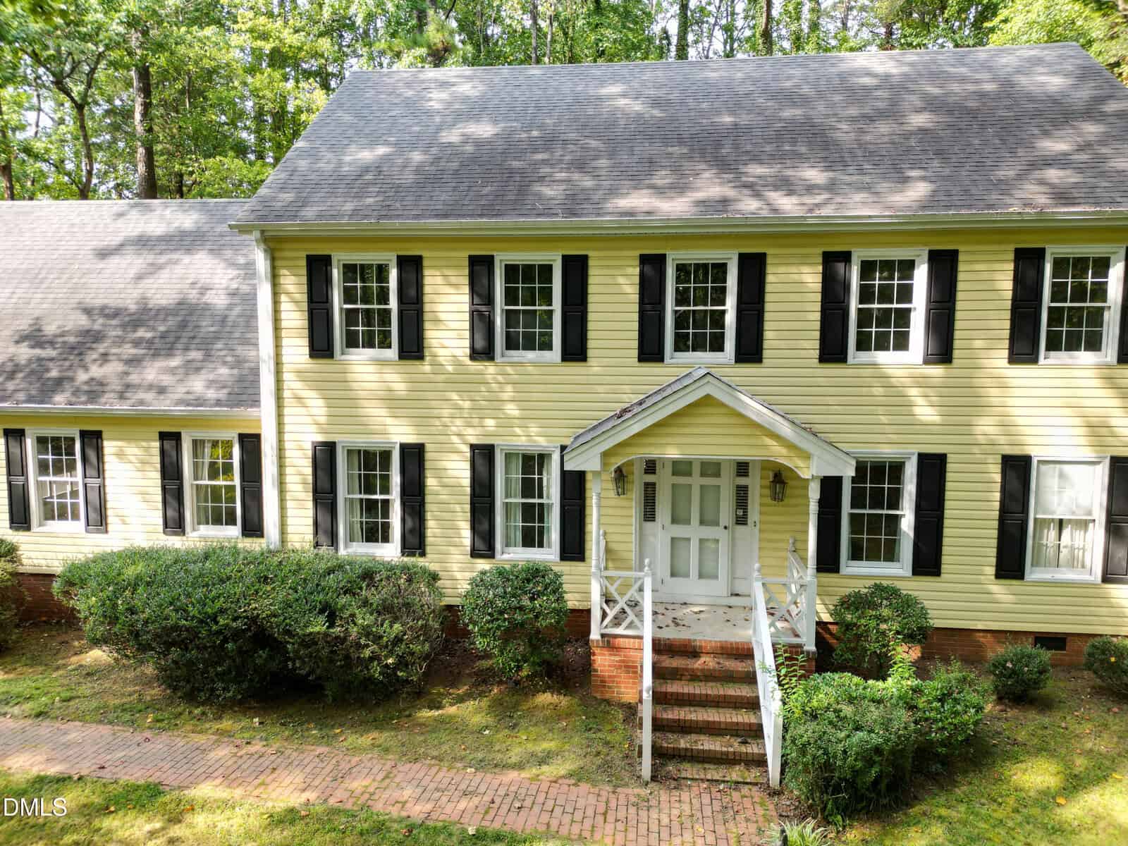 Two-story yellow house with black shutters, white trim, and a covered front porch with stairs. Brick walkway leads to the entrance. Shrubs and grass surround the house, and trees are visible in the background.