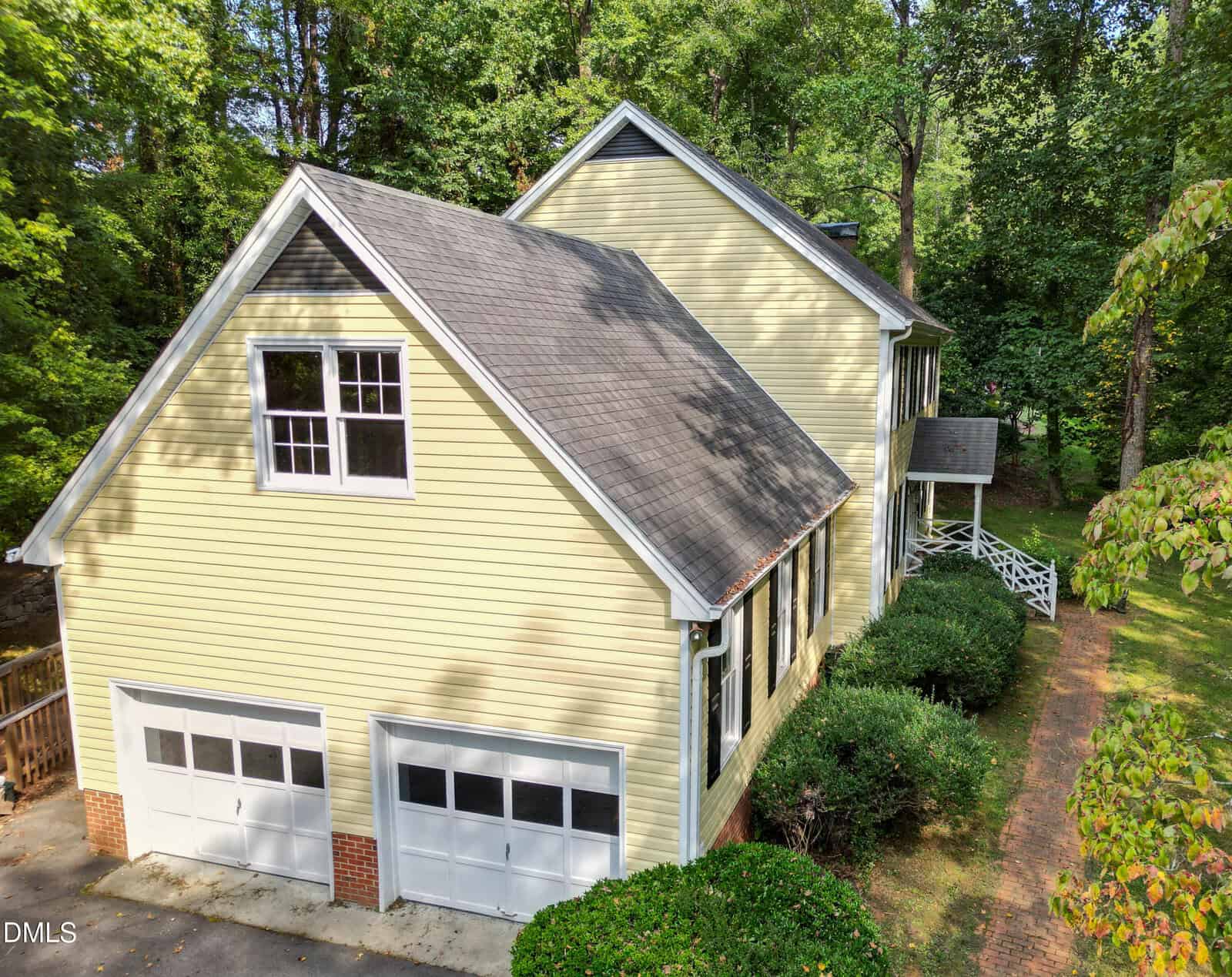 A two-story yellow house with white trim features a double garage, multiple windows, and a brick walkway surrounded by green trees and shrubs. The home is set in a wooded area.
