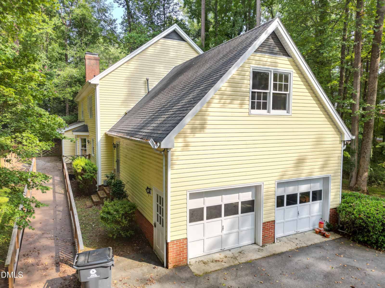 A two-story yellow house with white trim, double garage doors, and a steep roof, surrounded by trees. A concrete driveway and walkway lead to the side of the house, with a trash bin near the garage.