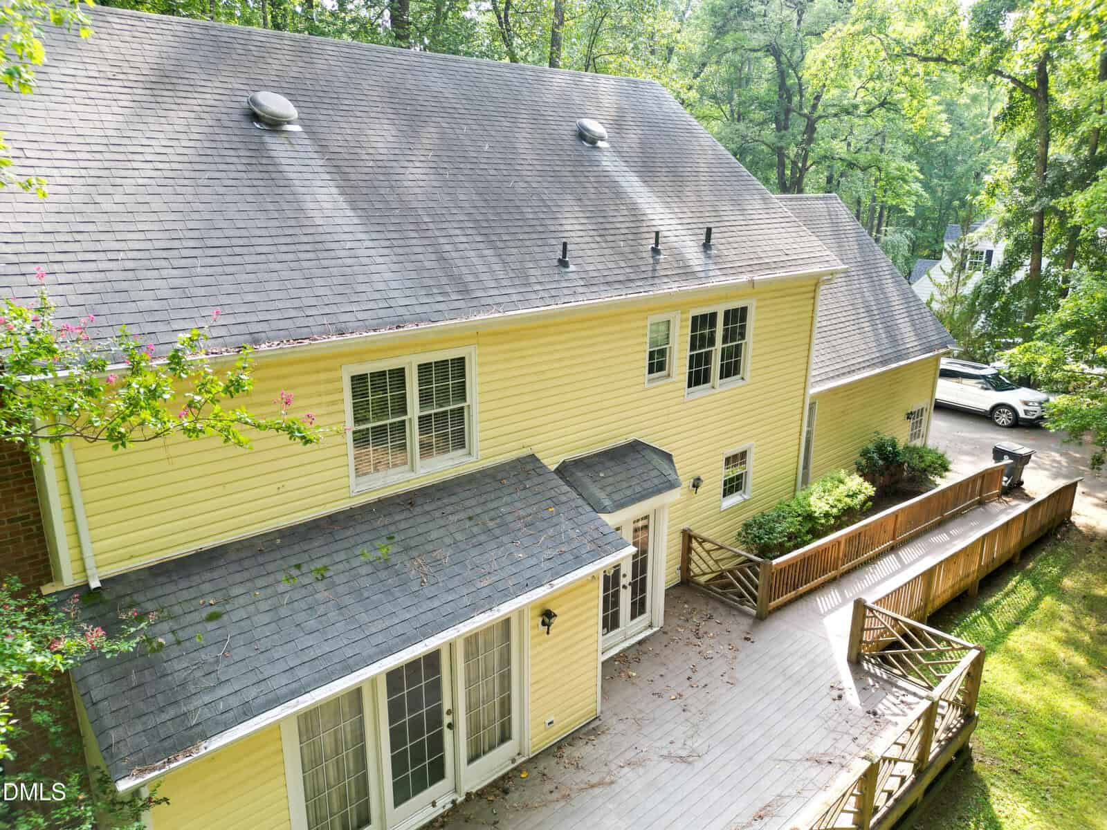 Aerial view of the back of a two-story yellow house with a gray shingled roof, a large wooden deck, multiple windows, and a driveway leading to a white parked vehicle surrounded by trees.