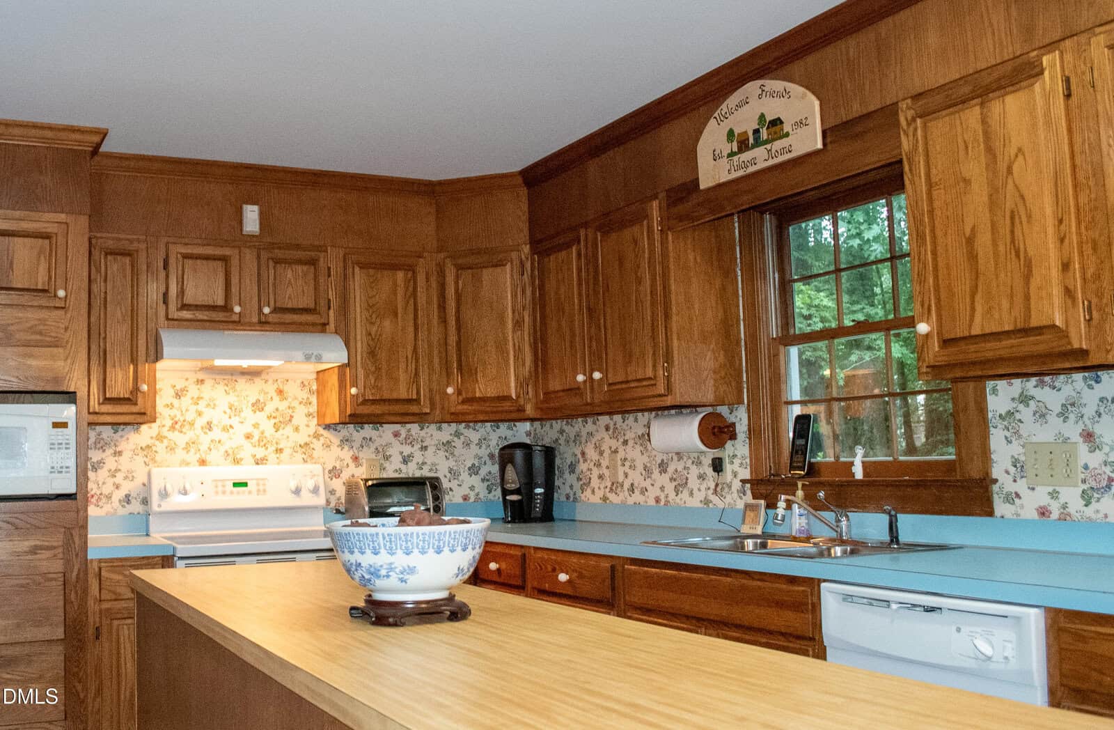 A kitchen with wood cabinets, a light blue countertop, floral wallpaper, and a large window over the sink. Appliances include a white stove with range hood, microwave, dishwasher, and a blue-and-white bowl on the island.