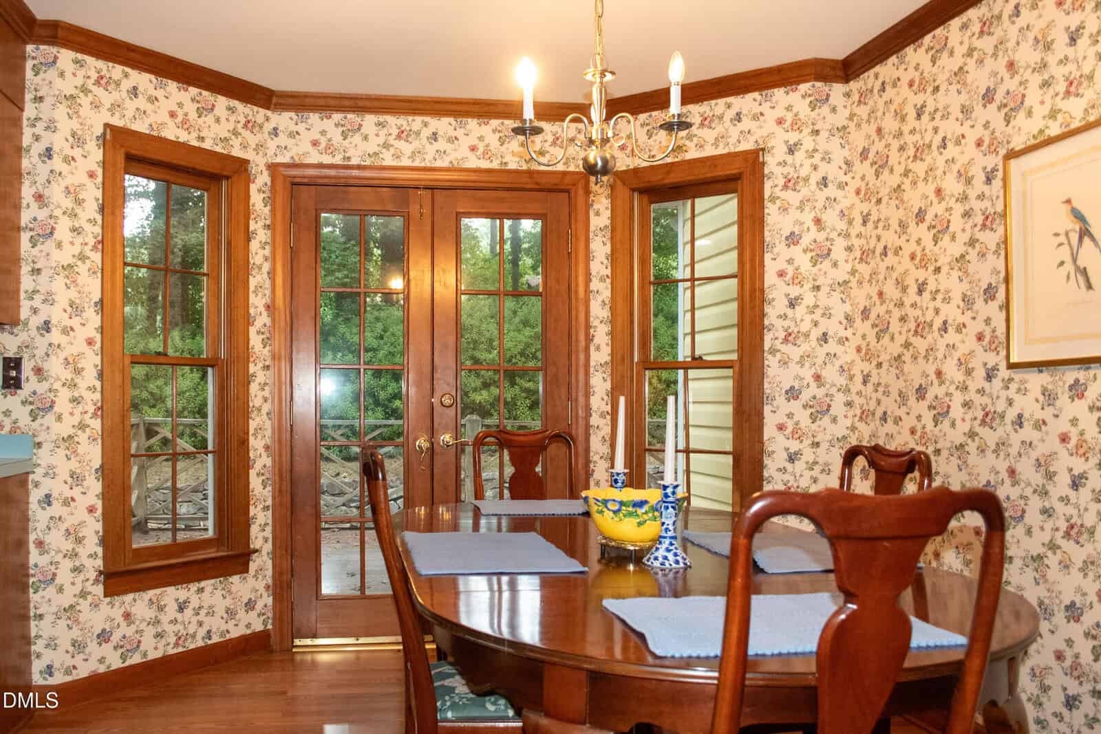 A dining room with floral wallpaper, a wooden table set with placemats, candles, and a yellow bowl, surrounded by wooden chairs. French doors and windows show a view of greenery outside.