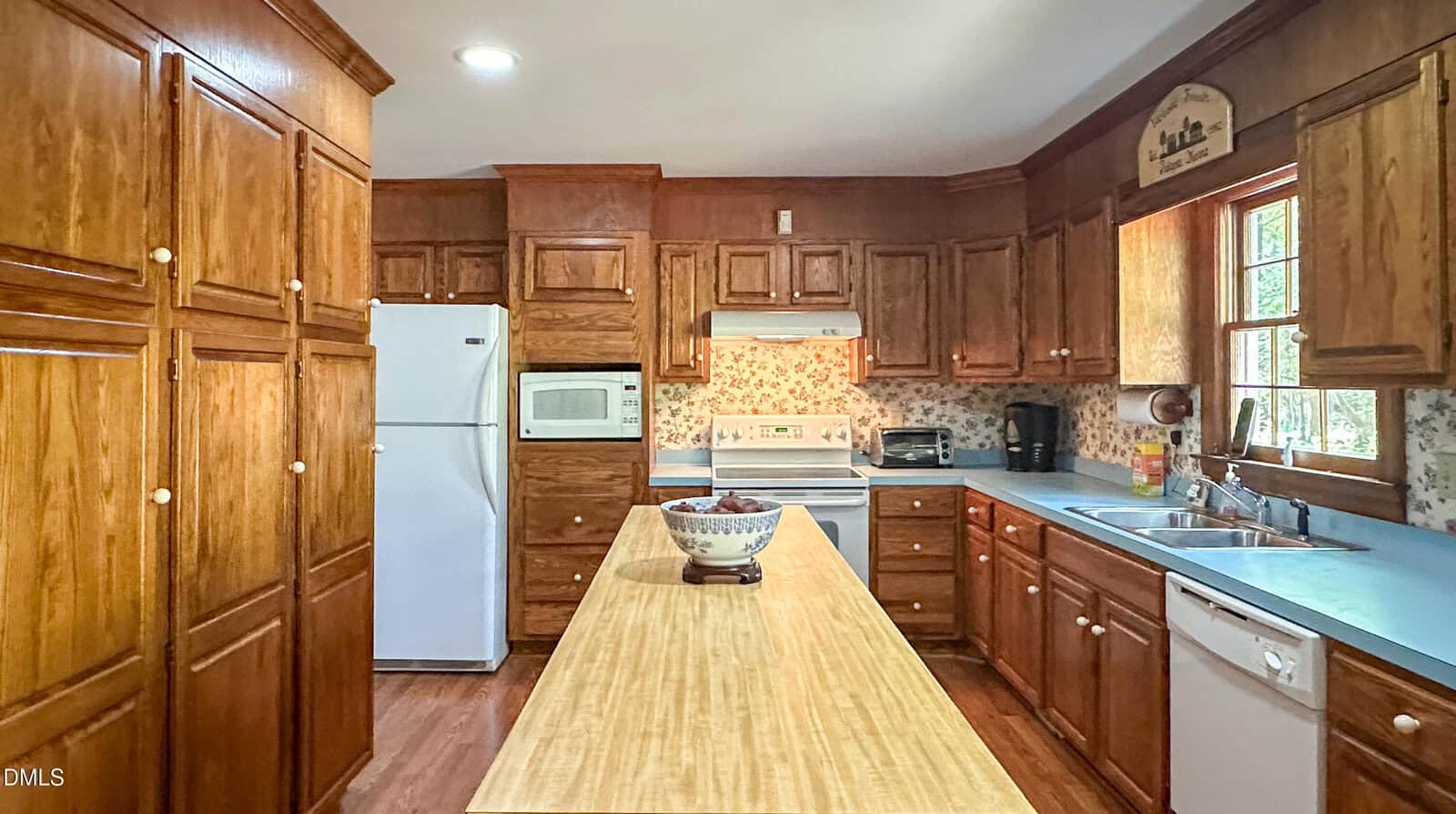 Spacious kitchen with wooden cabinets, white appliances, a central island with a bowl, blue countertops, a window above the sink, and floral wallpaper.