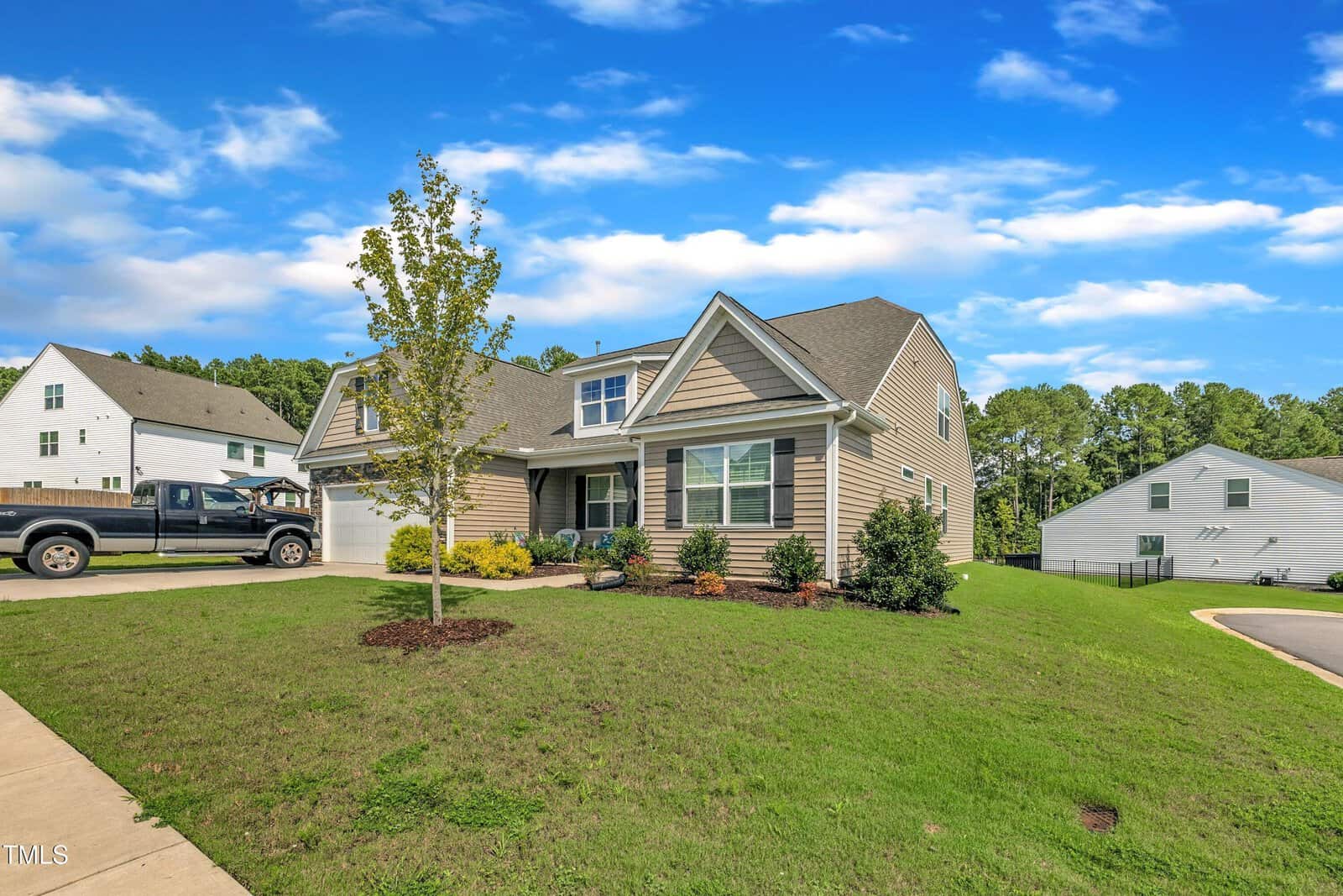 A two-story suburban house with tan siding, white trim, and a front-facing garage sits on a grassy lawn, with a young tree and landscaped plants by the entrance. A pickup truck is parked in the driveway.